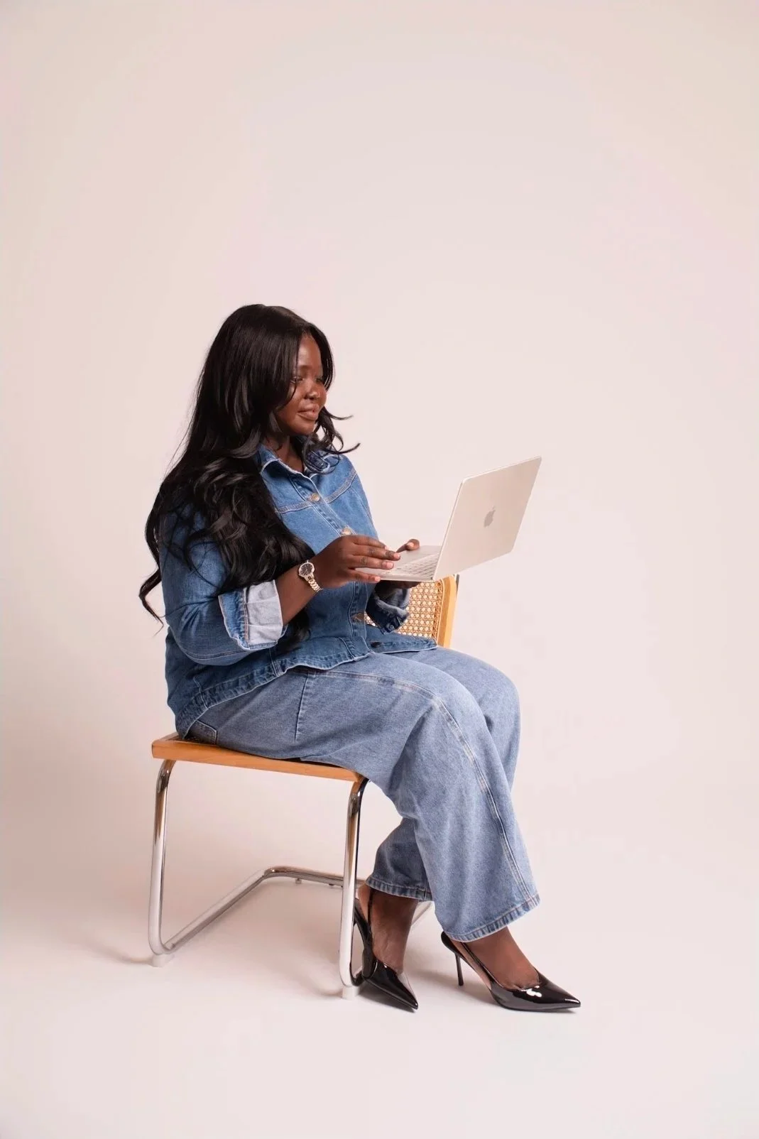 Woman sitting on a chair with a laptop, wearing a denim jacket, jeans, and high heels, against a plain background.