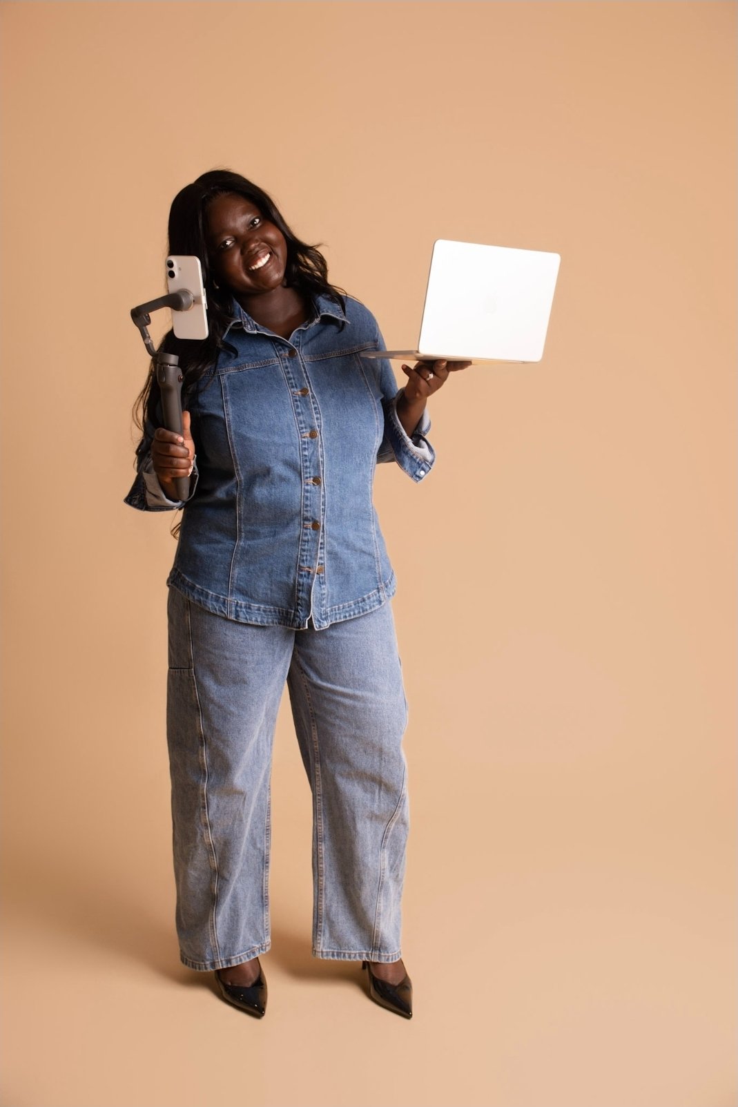 A woman holding a selfie stick with a phone, a laptop in her other hand, smiling and dressed in a denim shirt and jeans, standing against a plain beige background.