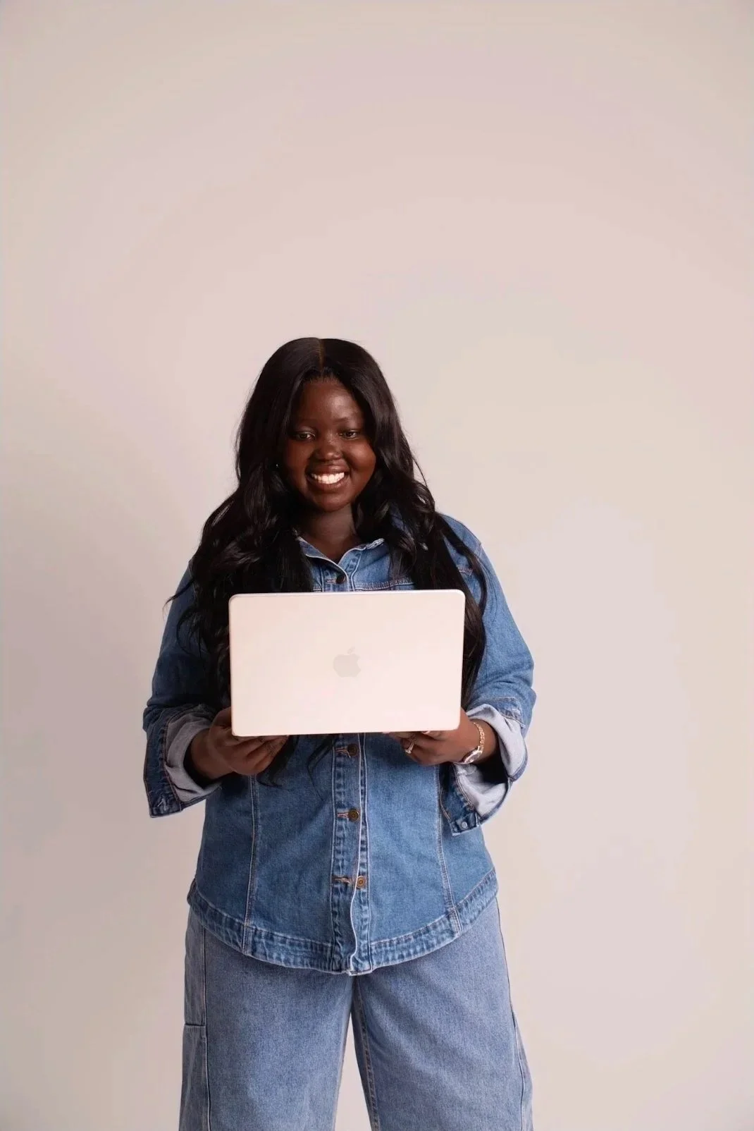 A woman with long black hair wearing a denim jacket and jeans, holding a silver Apple MacBook, and smiling while standing against a white background.