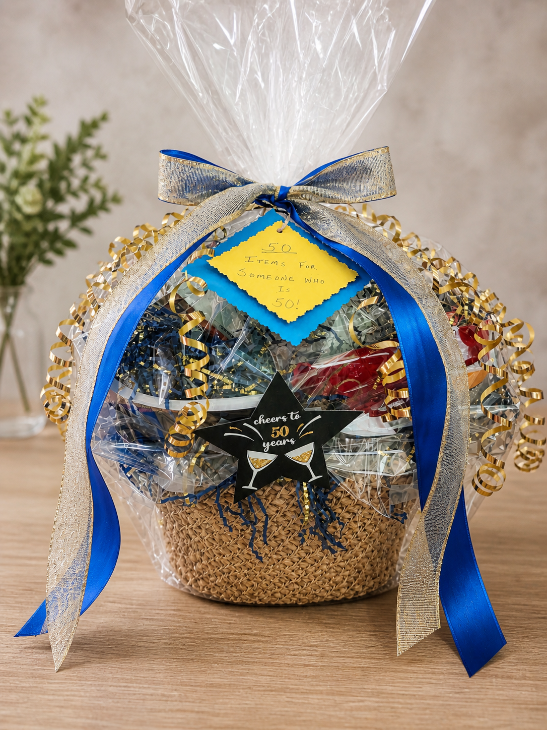 Gift basket decorated with gold and blue ribbons, containing 50 gifts for turning 50 and topped with a note celebrating a 50th birthday.