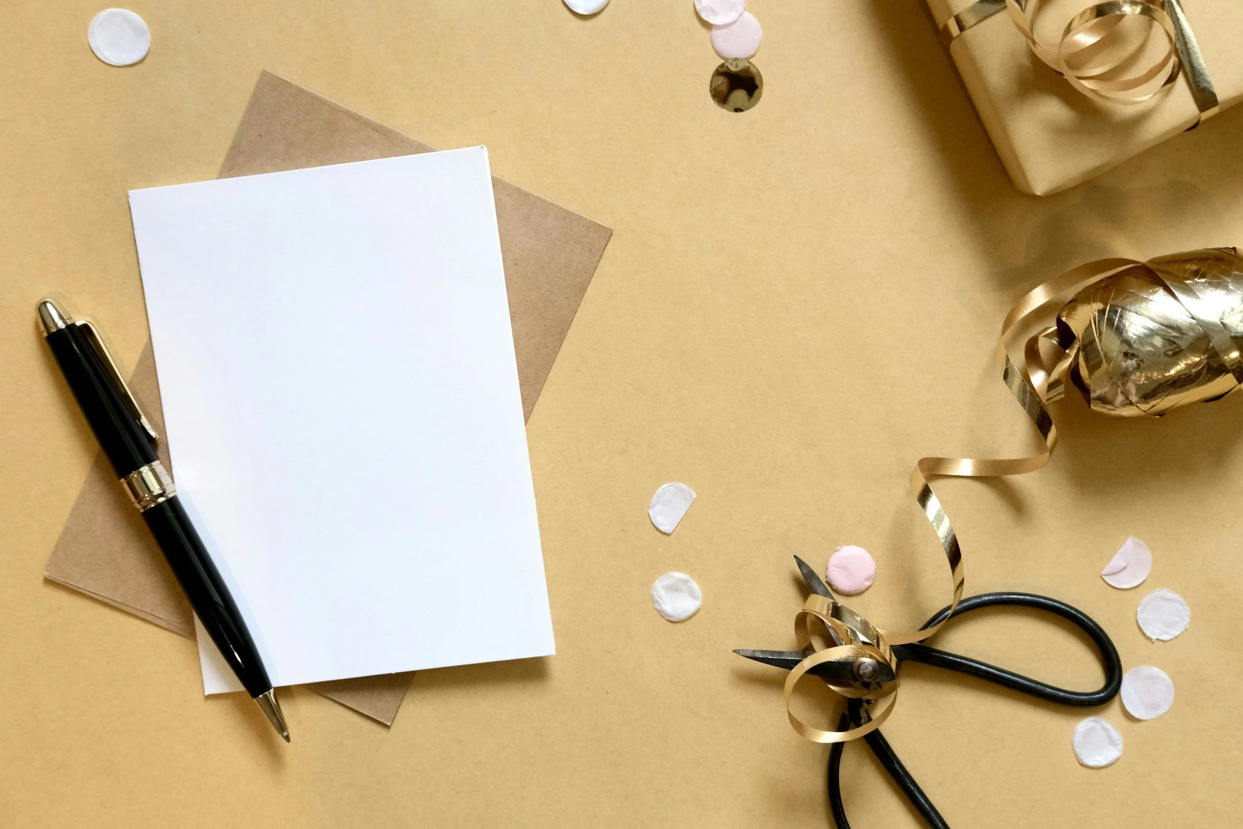 A celebration scene with a black pen, white and brown note cards, a pair of scissors, gold ribbon, a gold-wrapped balloon, and scattered pink and white confetti on a gold background.