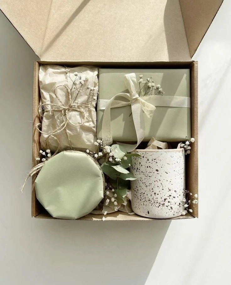 A gift box containing wrapped presents in white, green, and speckled designs, decorated with dried flowers and greenery.
