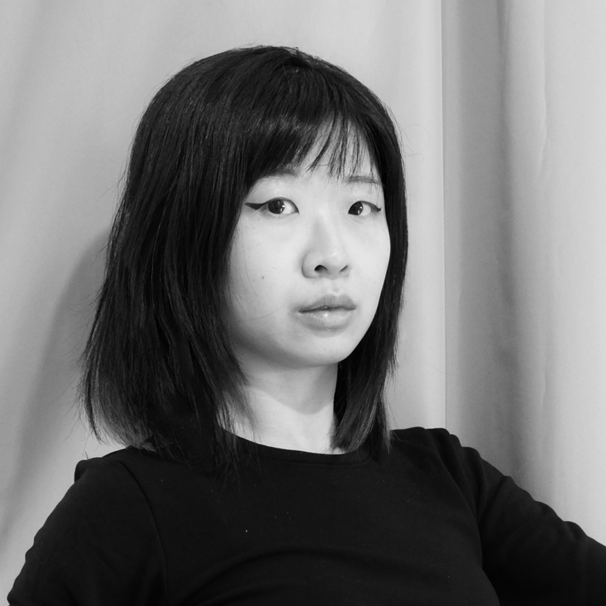 Black and white portrait of a woman with shoulder-length dark hair, wearing a dark top, standing against a plain background.