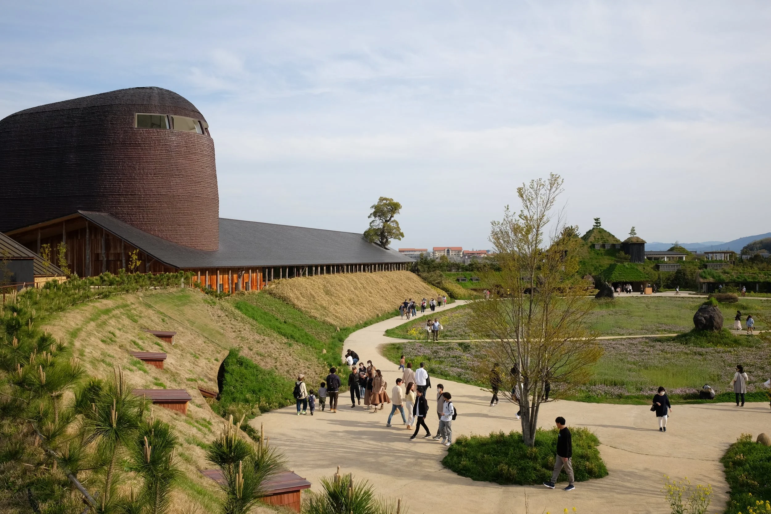 Visitors walking along a winding path through a landscaped park with traditional and modern buildings in the background, trees, and mountains in the distance.