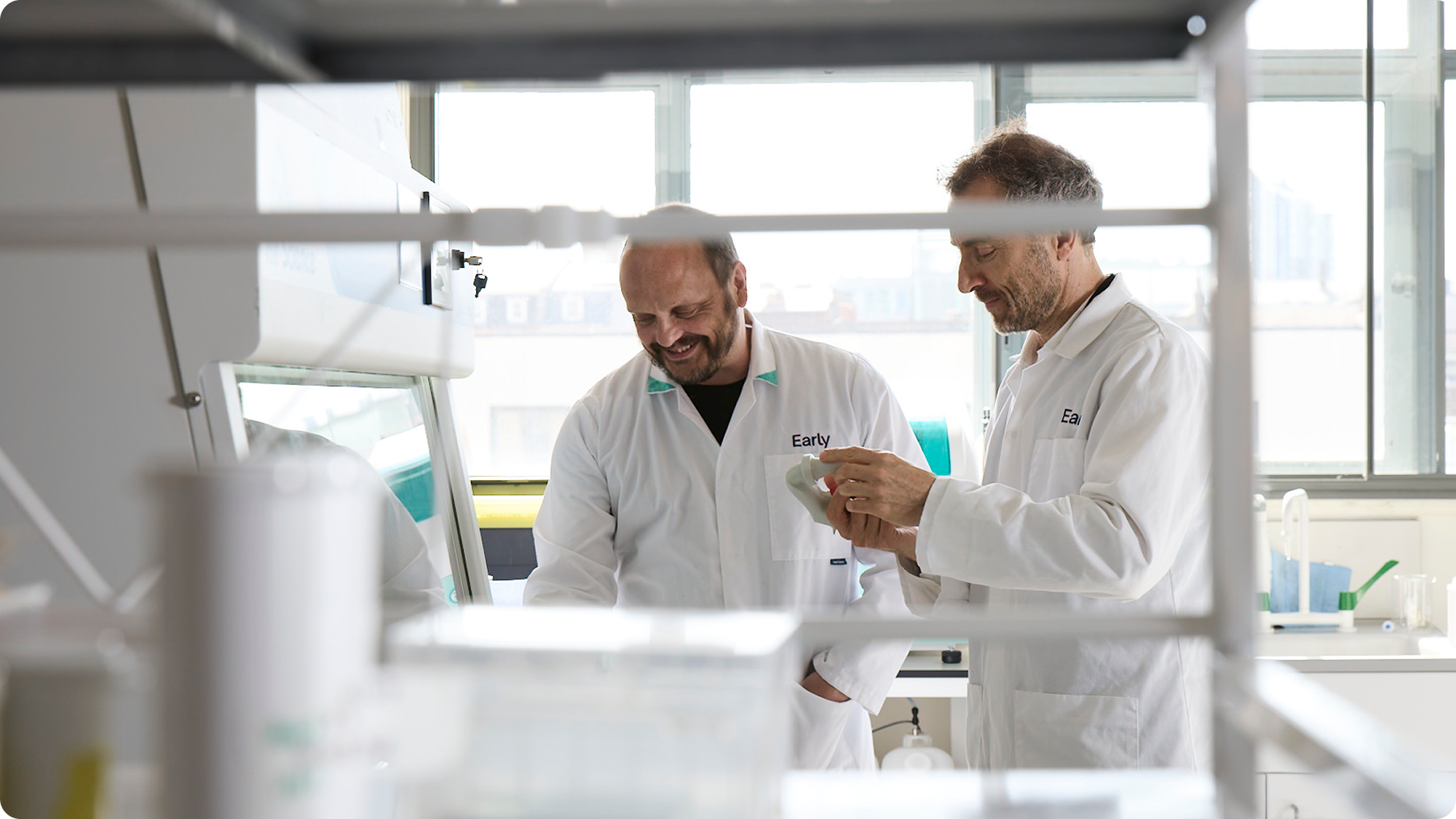 Two scientists in lab coats smiling and working together in a laboratory with lab equipment around them.