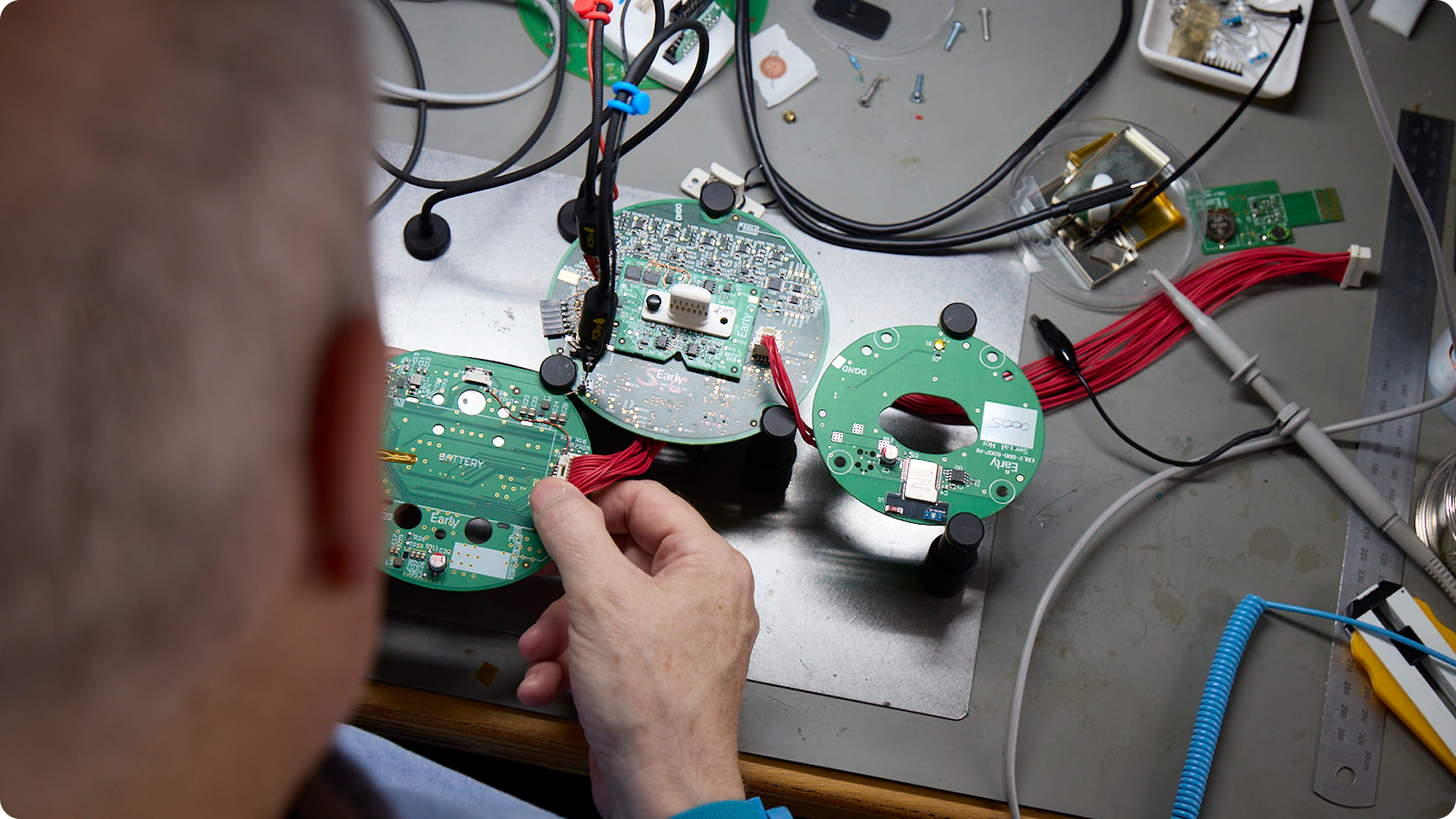 Person working on multiple green circuit boards and electronic components on a workbench surrounded by tools and wiring.