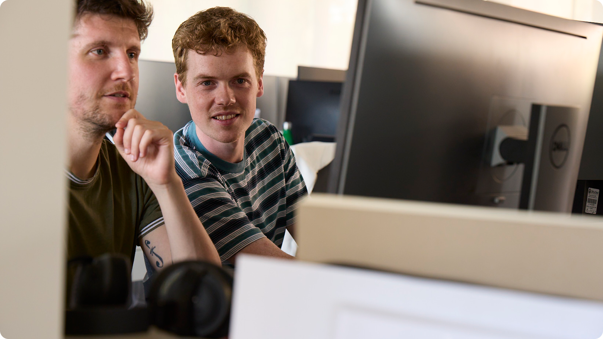 Two men sitting at a desk in front of computer monitors, engaged and smiling, in an office setting.