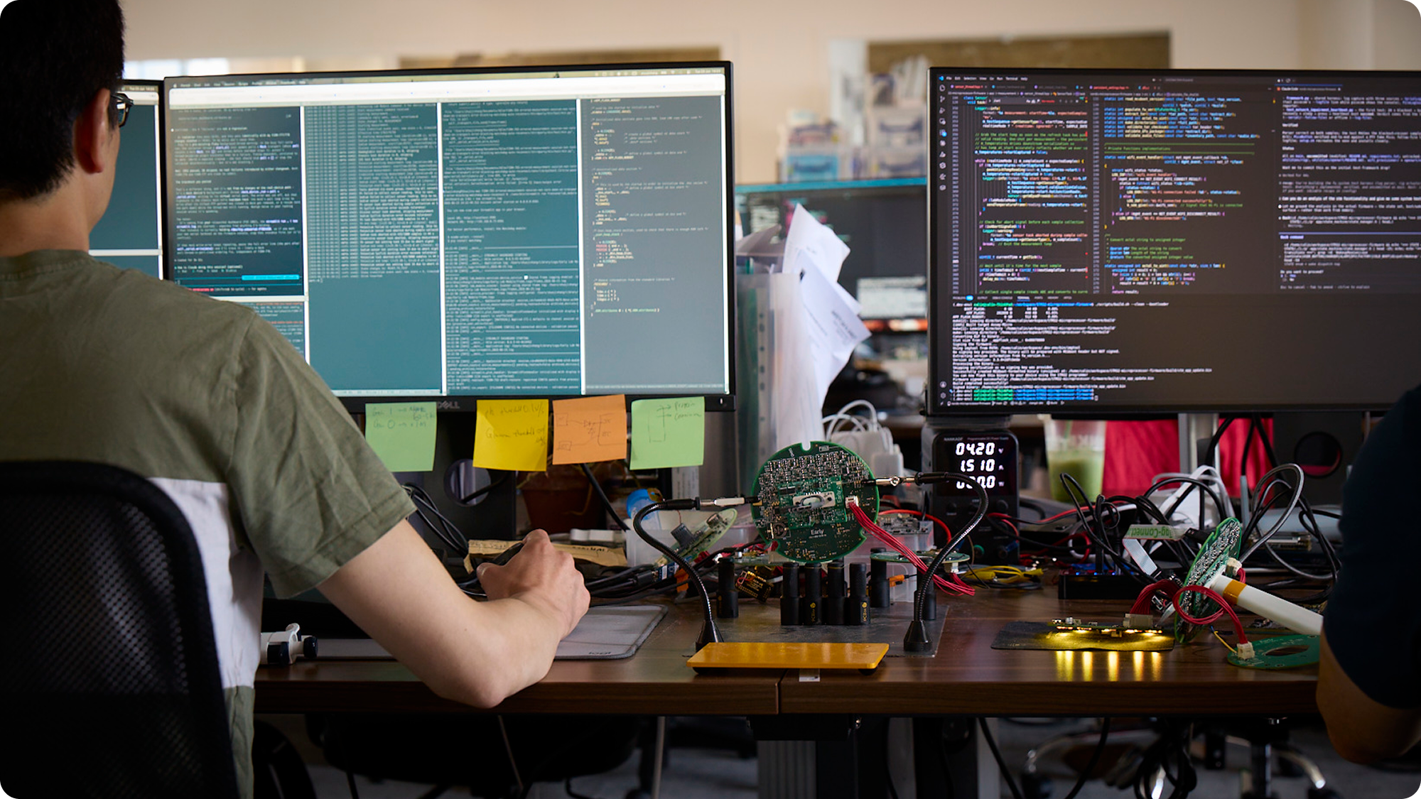 A person working at a desk with multiple computer monitors displaying lines of code, surrounded by electronic components, wires, and tools.