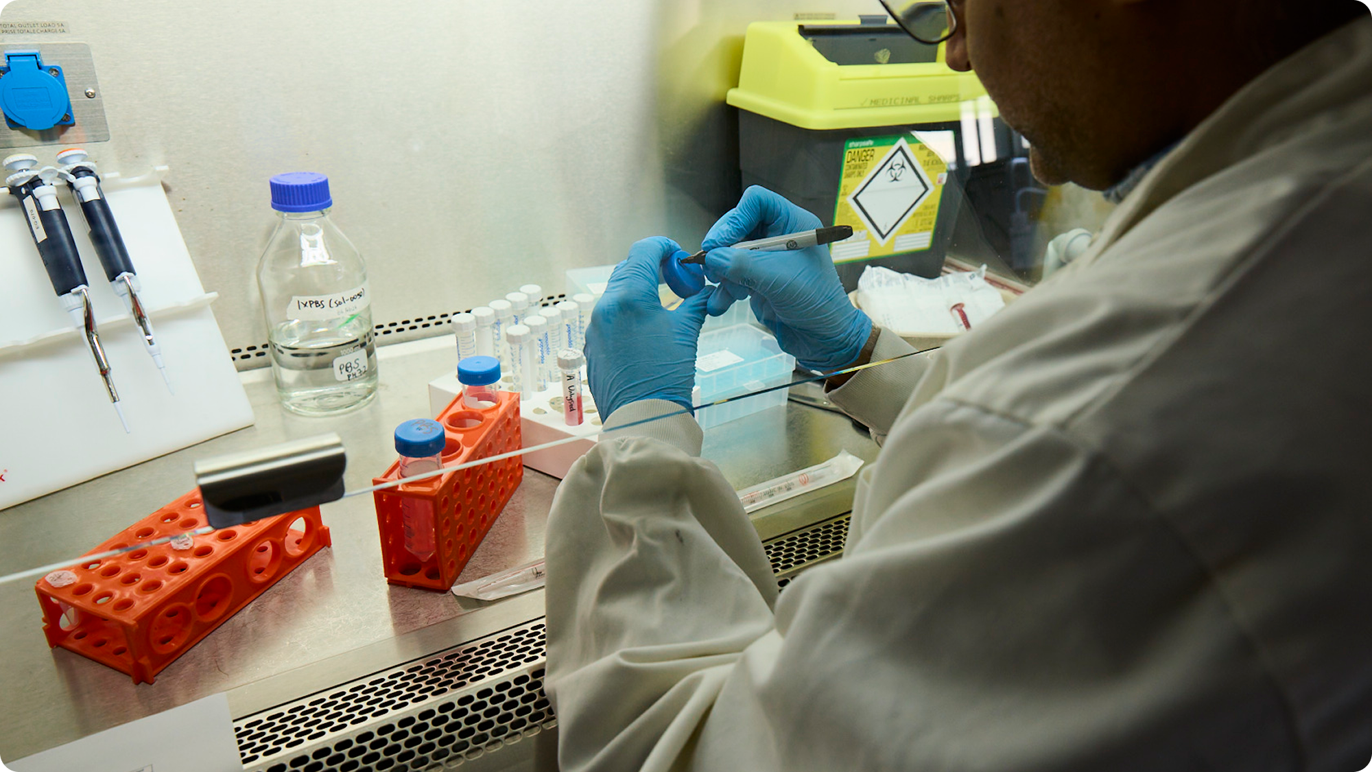 A scientist working in a laboratory, wearing blue gloves and using a pipette, surrounded by test tubes and laboratory equipment.