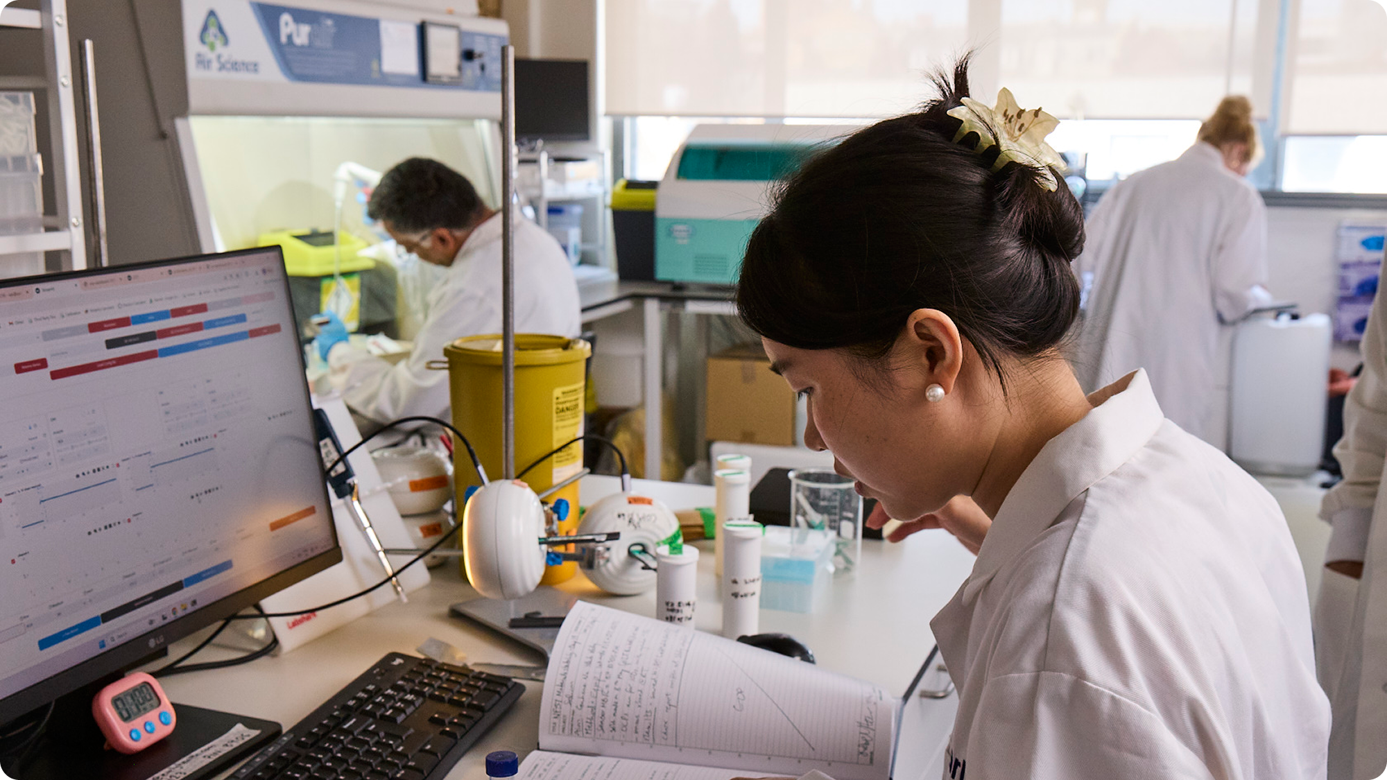Scientists in a lab working on experiments, with one scientist taking notes and a computer screen displaying data.
