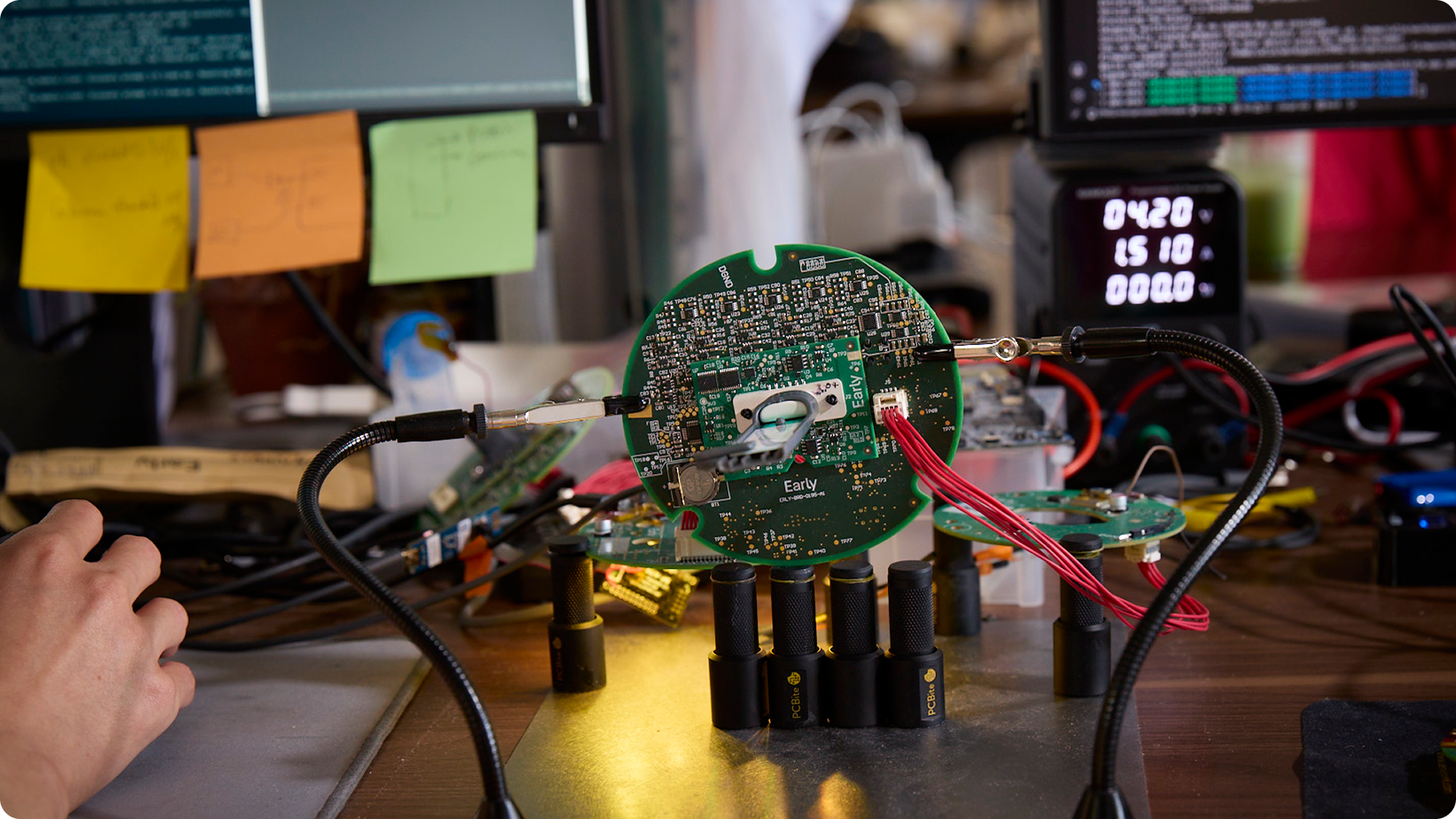 Close-up of a workbench with electronic components, circuit boards, and tools for hardware development, with multiple computer monitors and sticky notes in the background.
