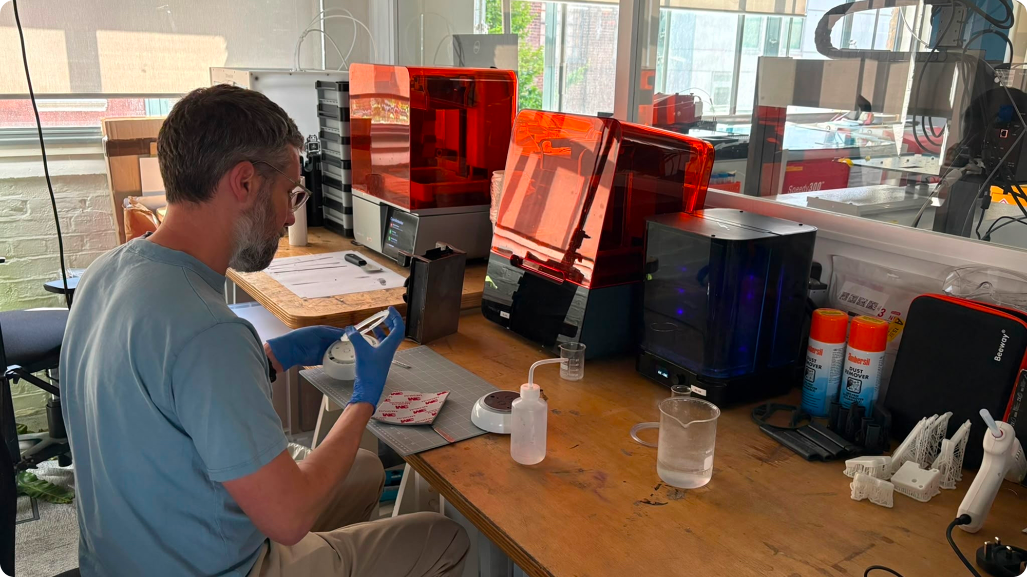 A man working on a electronic project at a desk with 3D printers, tools, and supplies