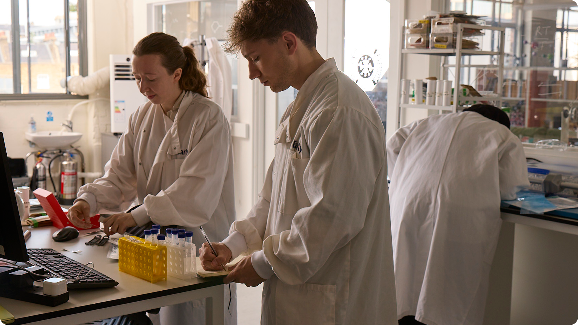Scientists working in a laboratory, wearing white lab coats. One woman and one man are concentrating on their tasks near a desk with lab equipment, test tubes, and a computer.