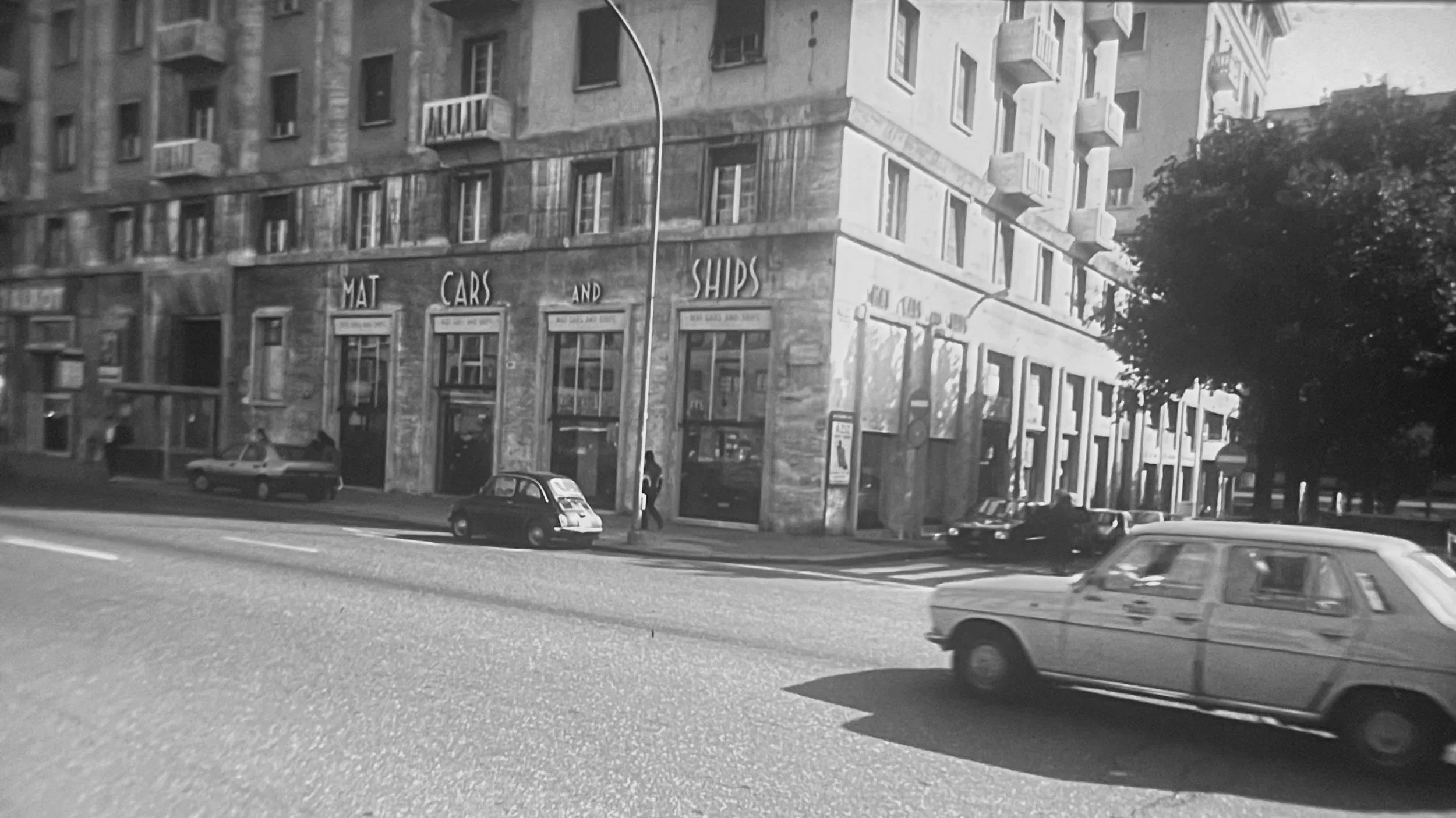 Black and white photo of a city street with cars and pedestrians, including a building with signs reading 'MAT CARS AND SHIPS' and several buildings with multiple floors and balconies.