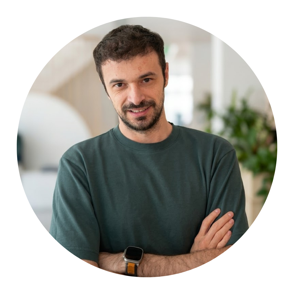 A man with dark hair and a beard, wearing a dark green T-shirt and a smartwatch, stands with his arms crossed, smiling in a brightly lit indoor setting with plants in the background.