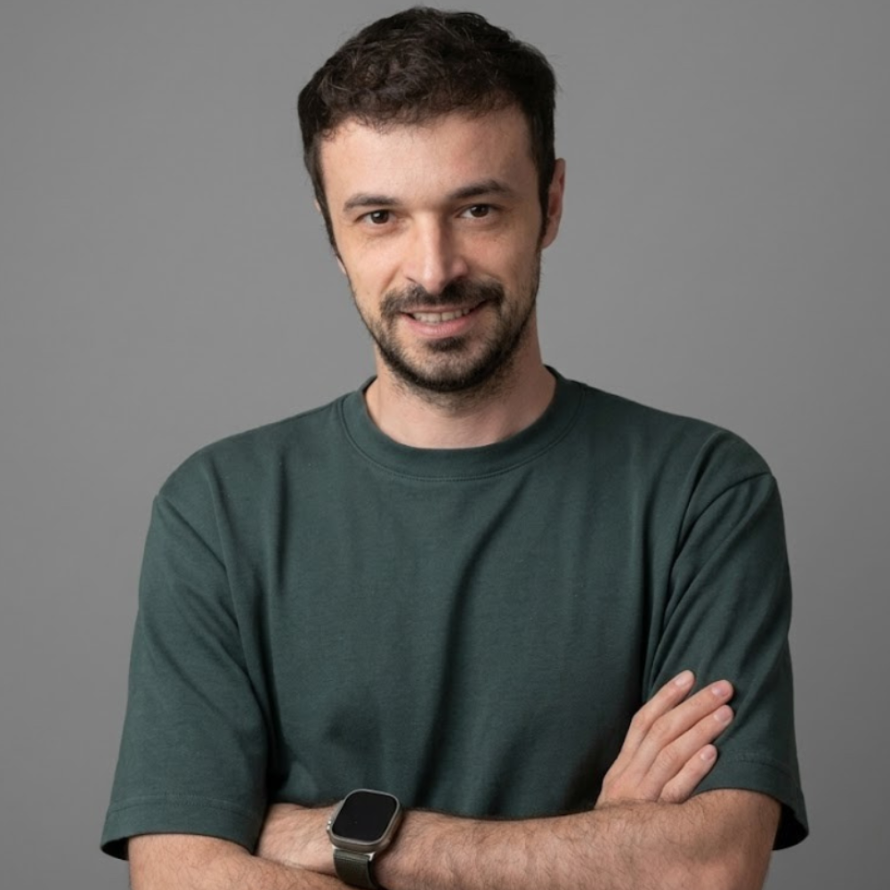 A man with short brown hair, a beard, and a mustache, smiling and crossing his arms, wearing a dark green t-shirt and a smartwatch on his left wrist, standing against a plain gray background.