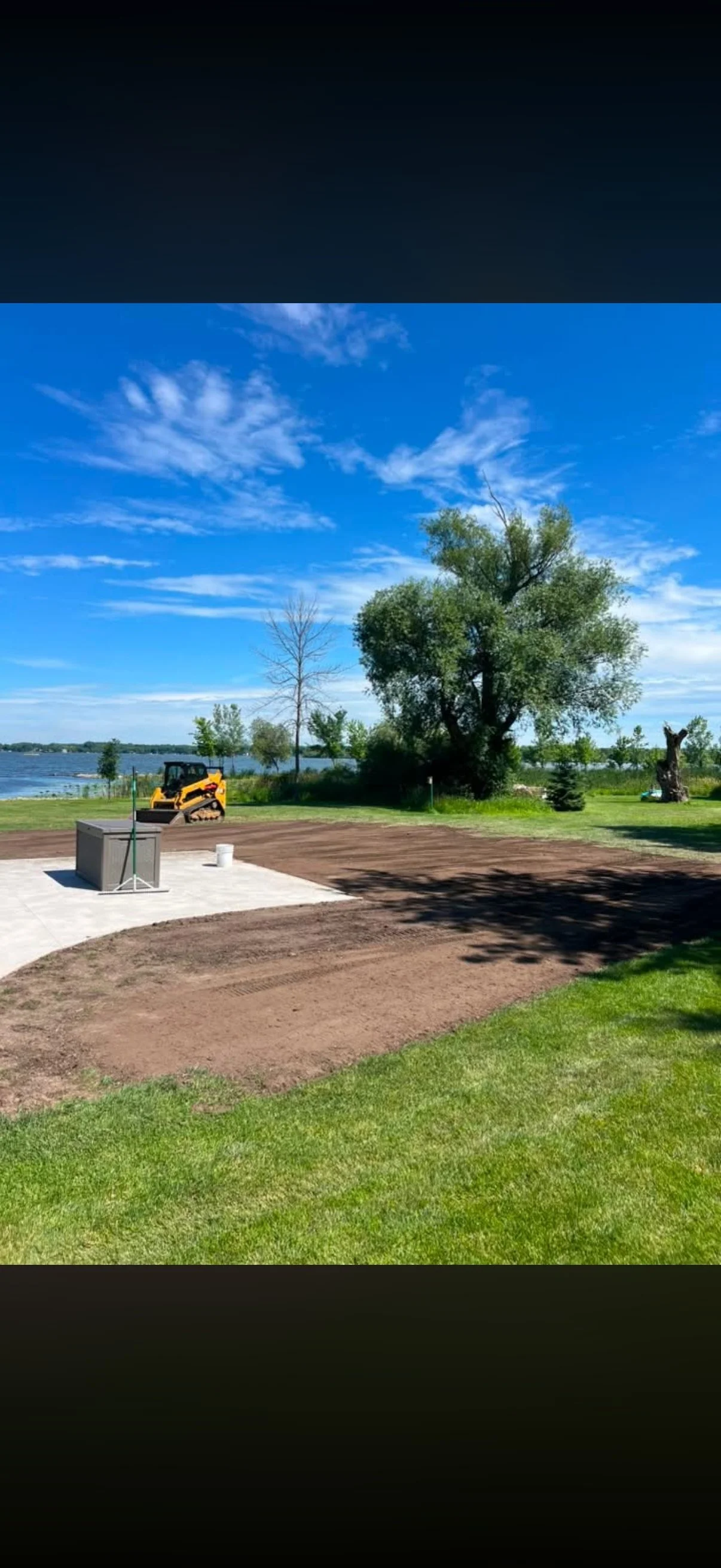 A lakeside park with a partially completed outdoor patio, a small construction vehicle, and a large leafy tree under a blue sky with scattered clouds.