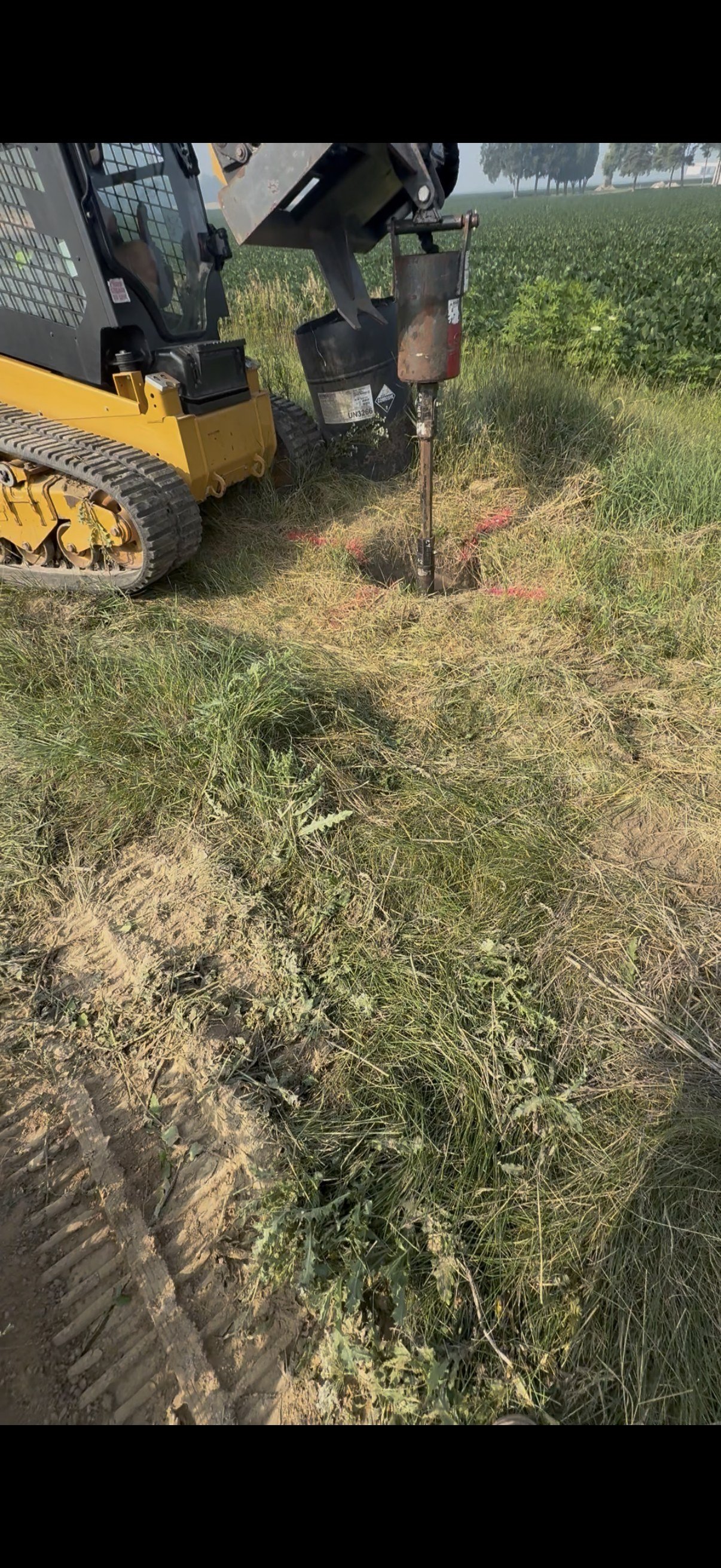 Close-up of a construction mini excavator digging a hole in grassy soil with a metal post and brown bucket attachment, in a field.
