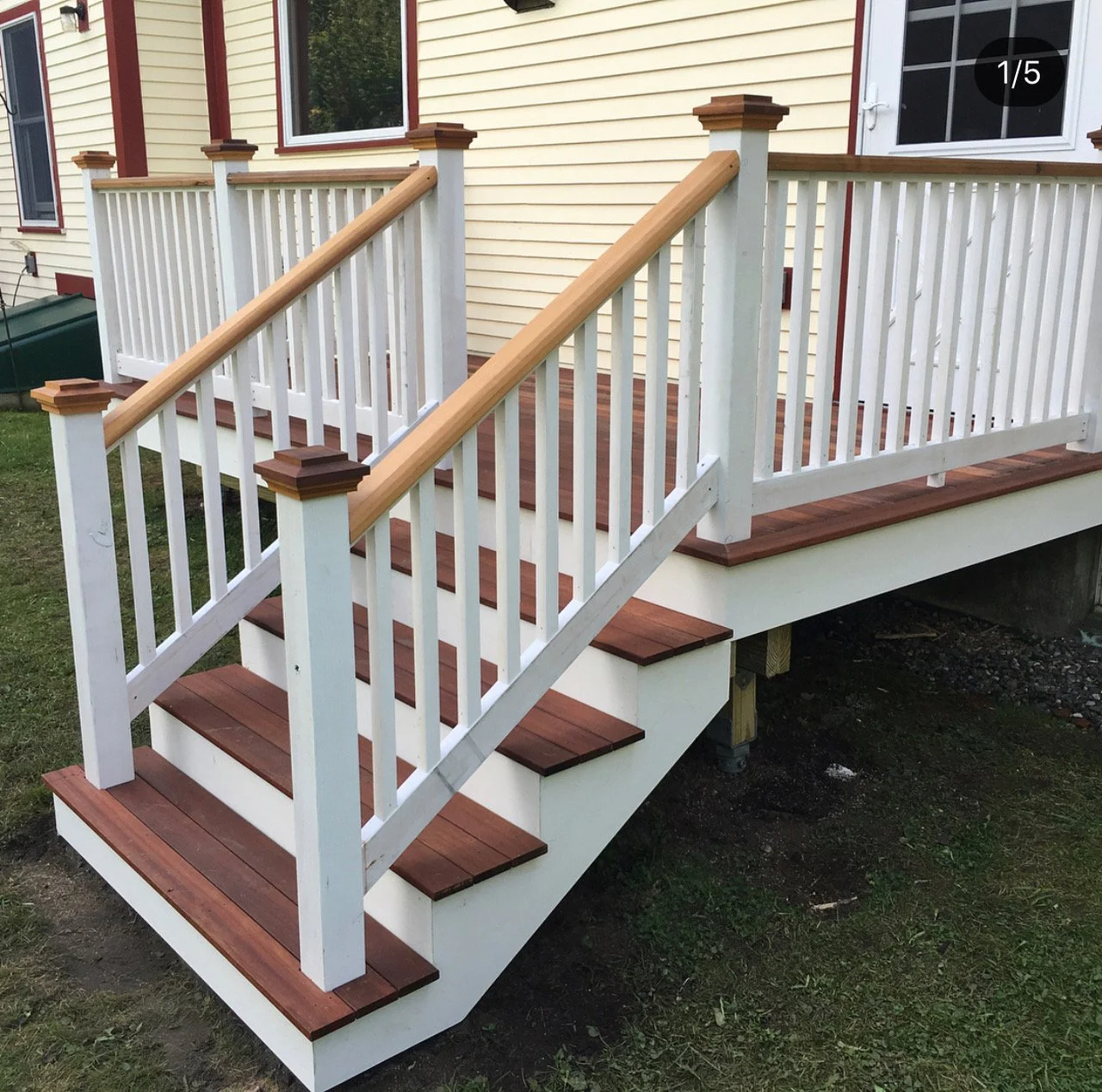 Wooden deck with white railing and stairs with brown steps in front of a house with yellow siding and white trim.