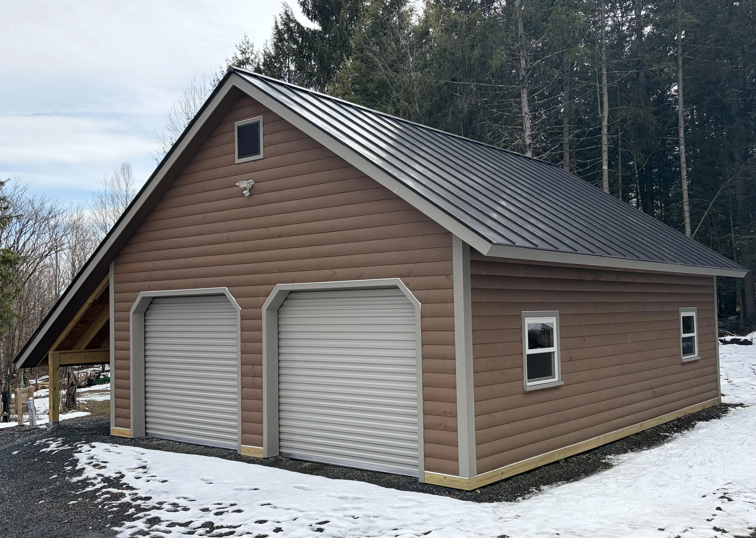 A new wooden garage with a metal roof, two roller doors, and a small window, surrounded by snow and tall trees in the background.