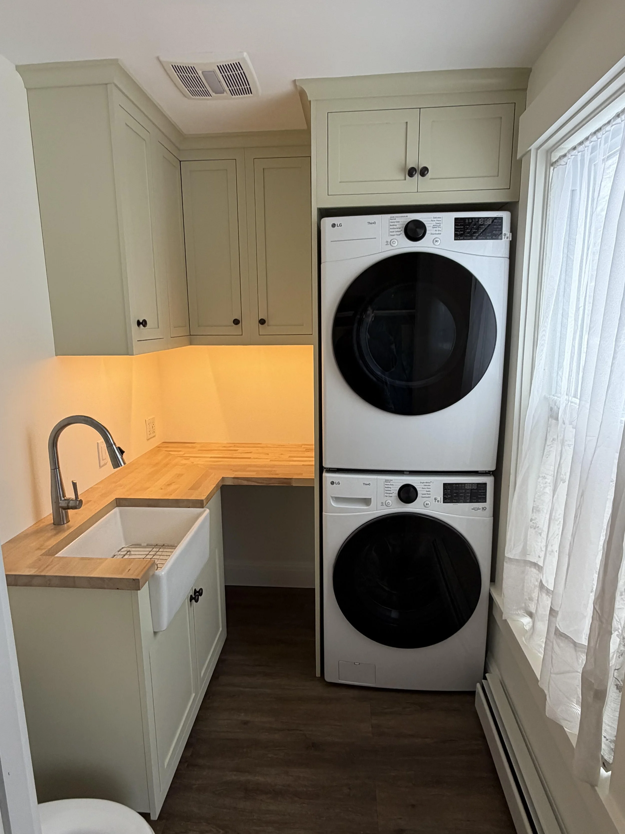 A laundry room with a stacked washer and dryer, a small white utility sink, cream-colored cabinets, a window with white curtains, and a dark wood floor.