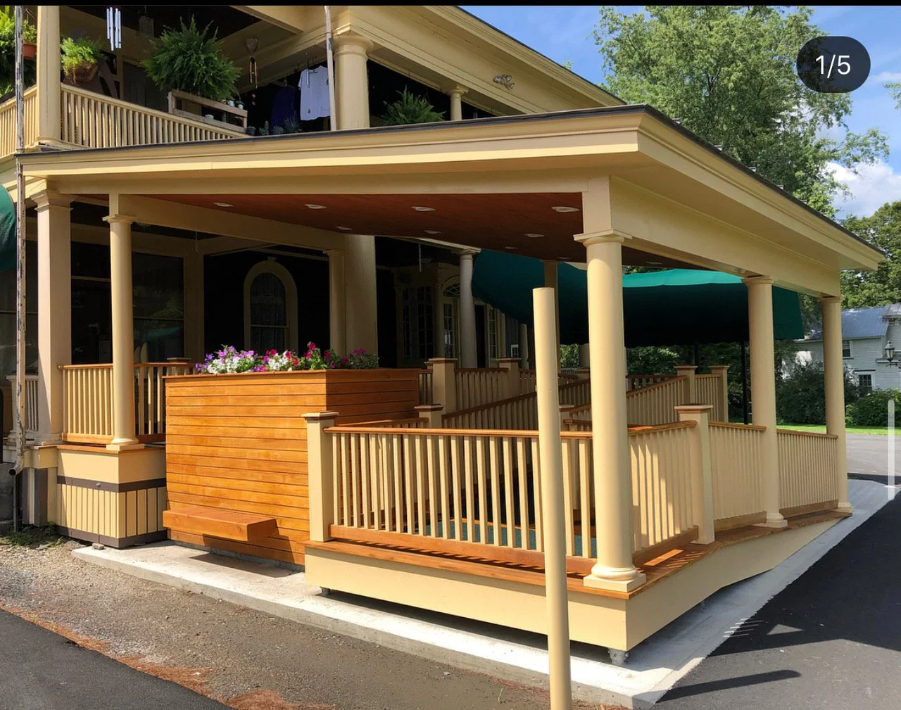 Front porch of a house with beige columns, wooden and beige railings, a wooden deck with flower boxes, green awning, and mature trees in the background.