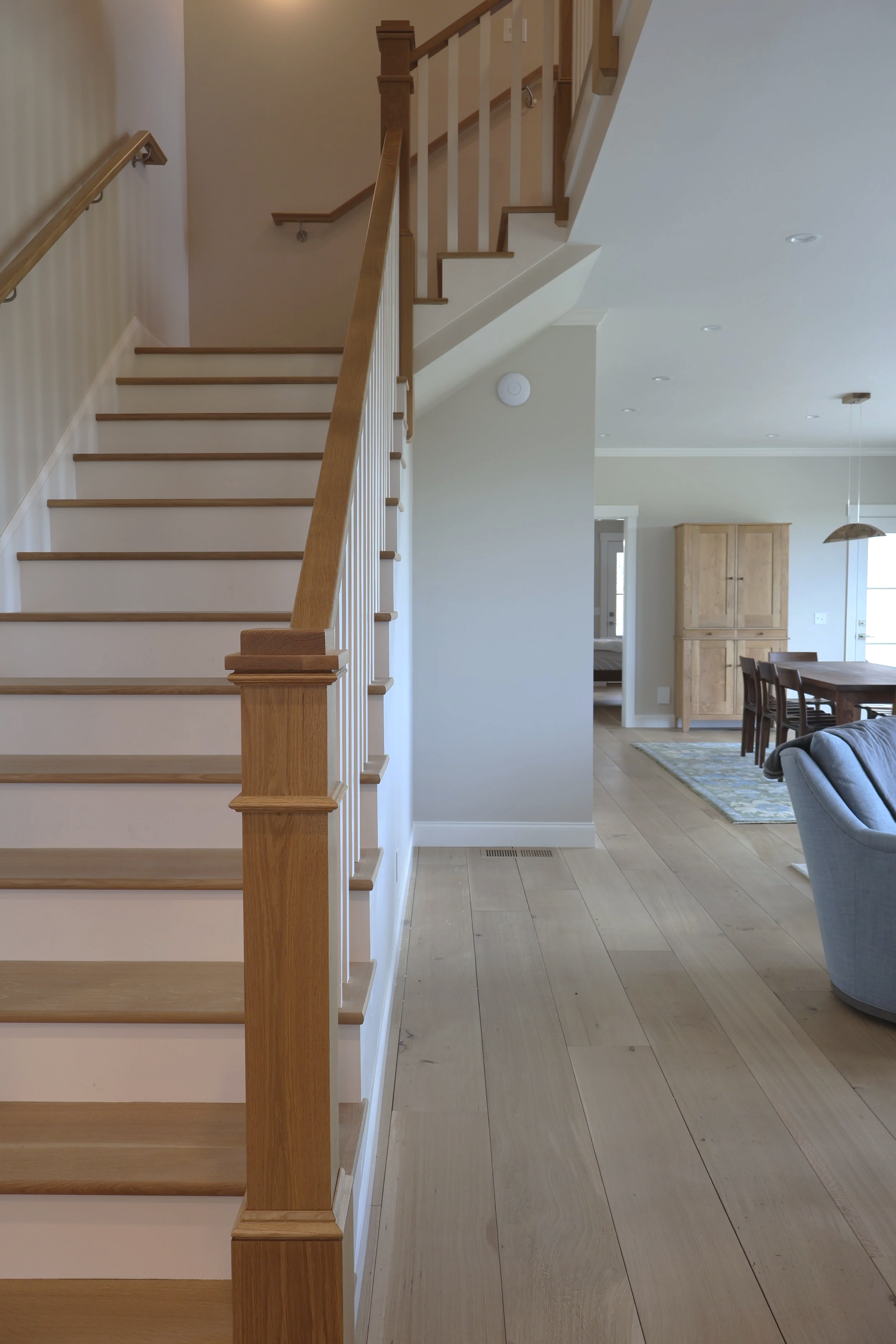 Inside of a modern home showing a staircase with wooden handrails and white balusters, a living area with a wooden dining table and chairs, a wooden cabinet, and a light-colored hardwood floor.