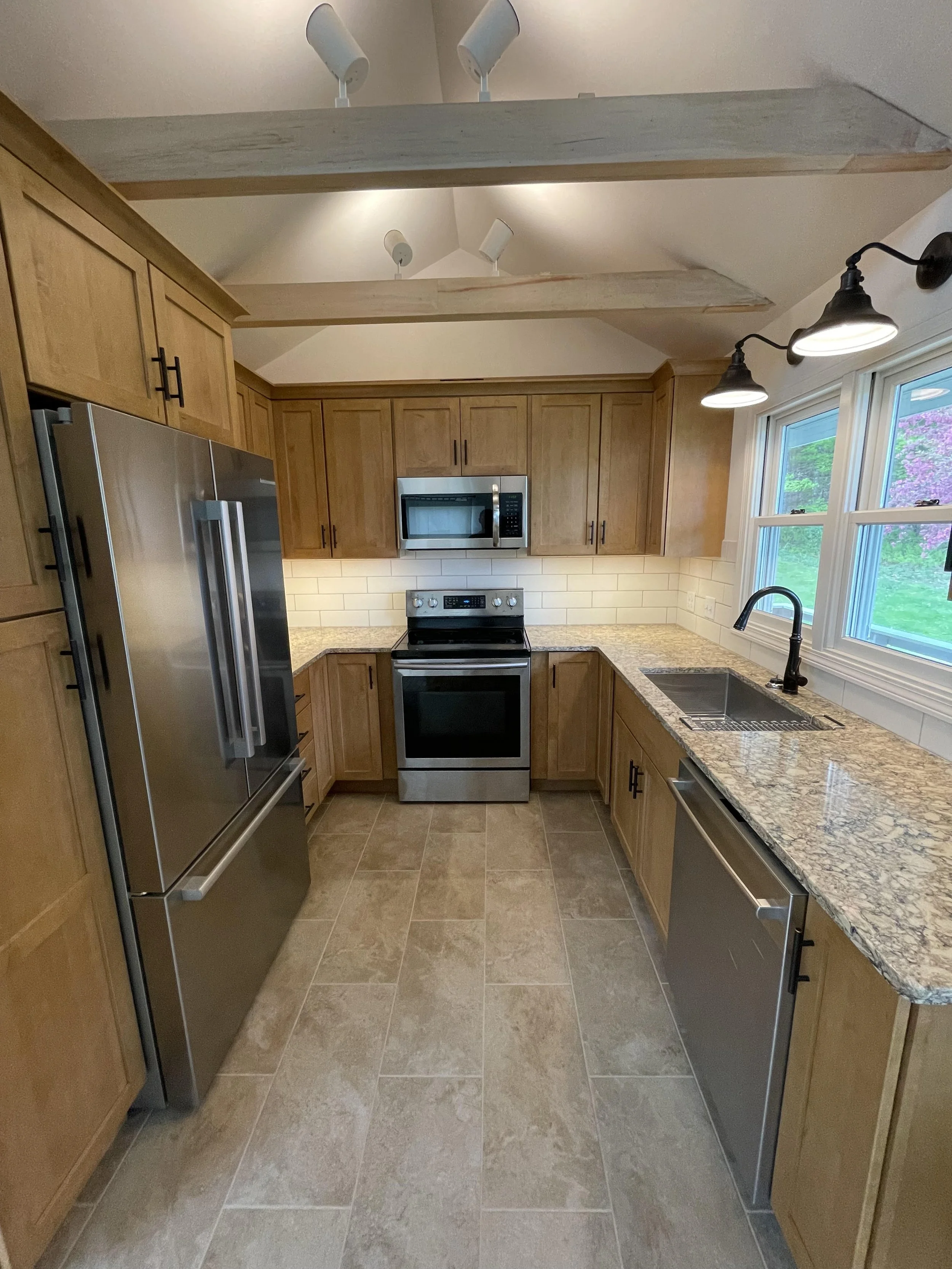 A modern kitchen with wooden cabinets, stainless steel refrigerator and stove, granite countertops, a double-basin sink with gooseneck faucet, tile flooring, and a window showing a view of greenery and purple flowers.