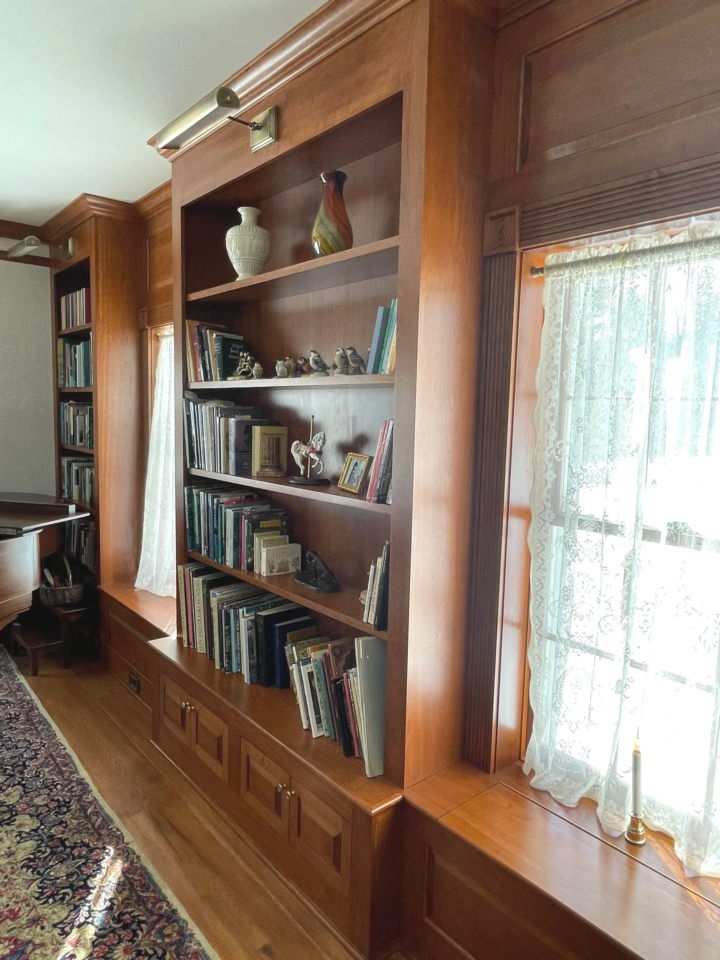 Wooden bookcase filled with books, decorative vases, and small figurines, next to a window with lace curtains in a room with a patterned rug.