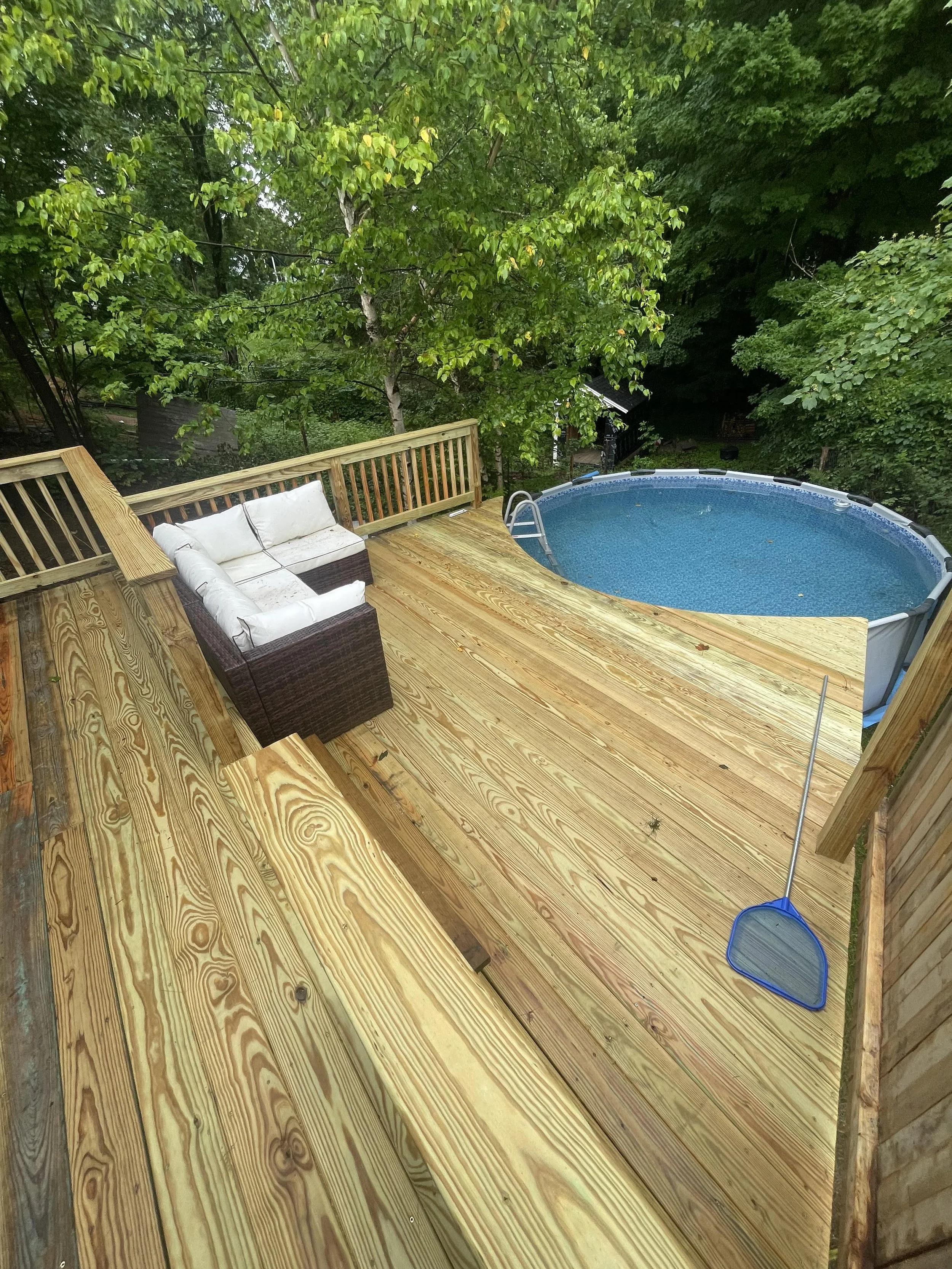 A backyard wooden deck with a swimming pool, outdoor furniture including a sofa with white cushions, surrounded by green trees and foliage.