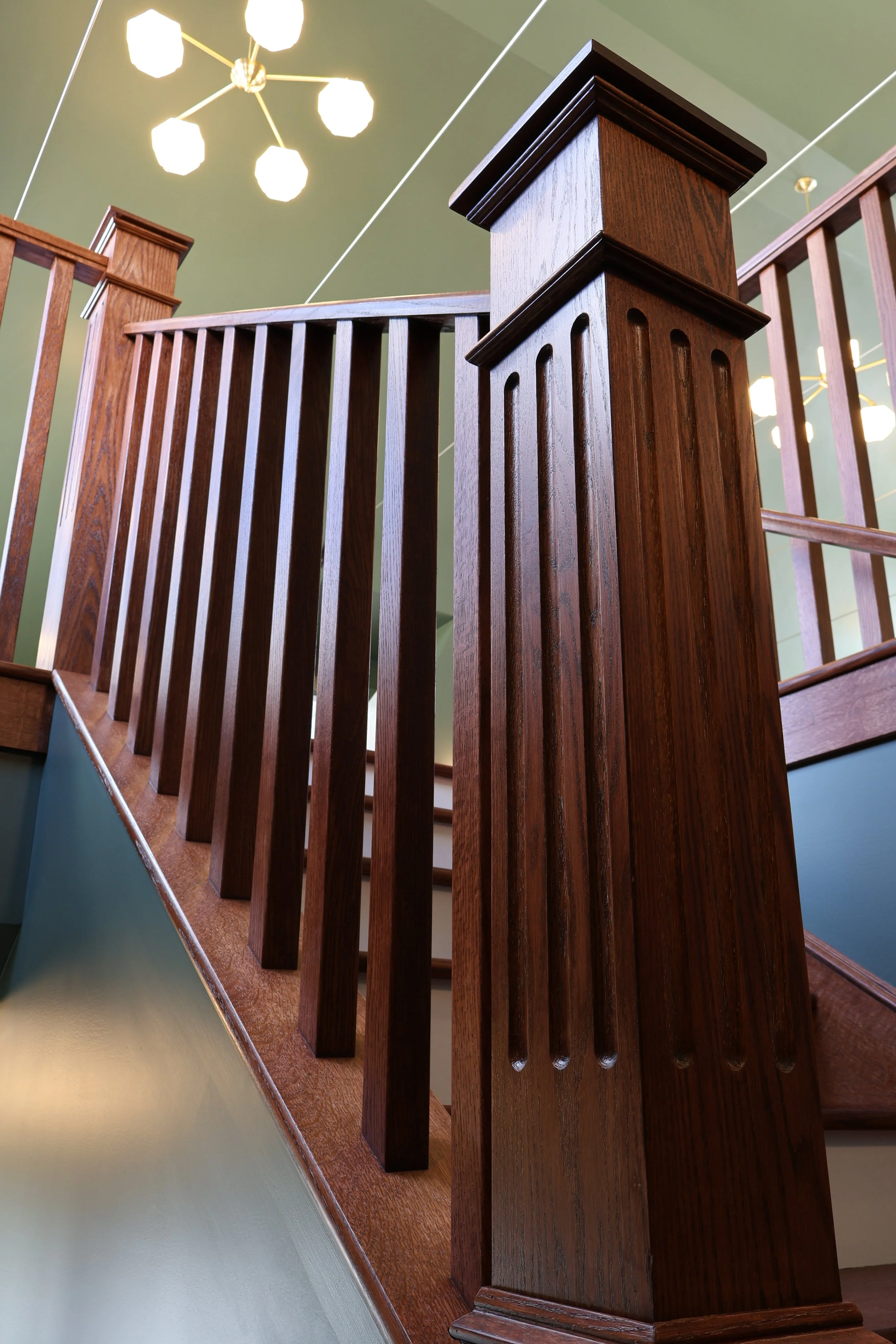 A close-up view of a wooden staircase railing and newel post, with a modern ceiling light fixture visible above. The staircase is made of polished dark wood, and the wall adjacent to the staircase is painted in a light muted color.