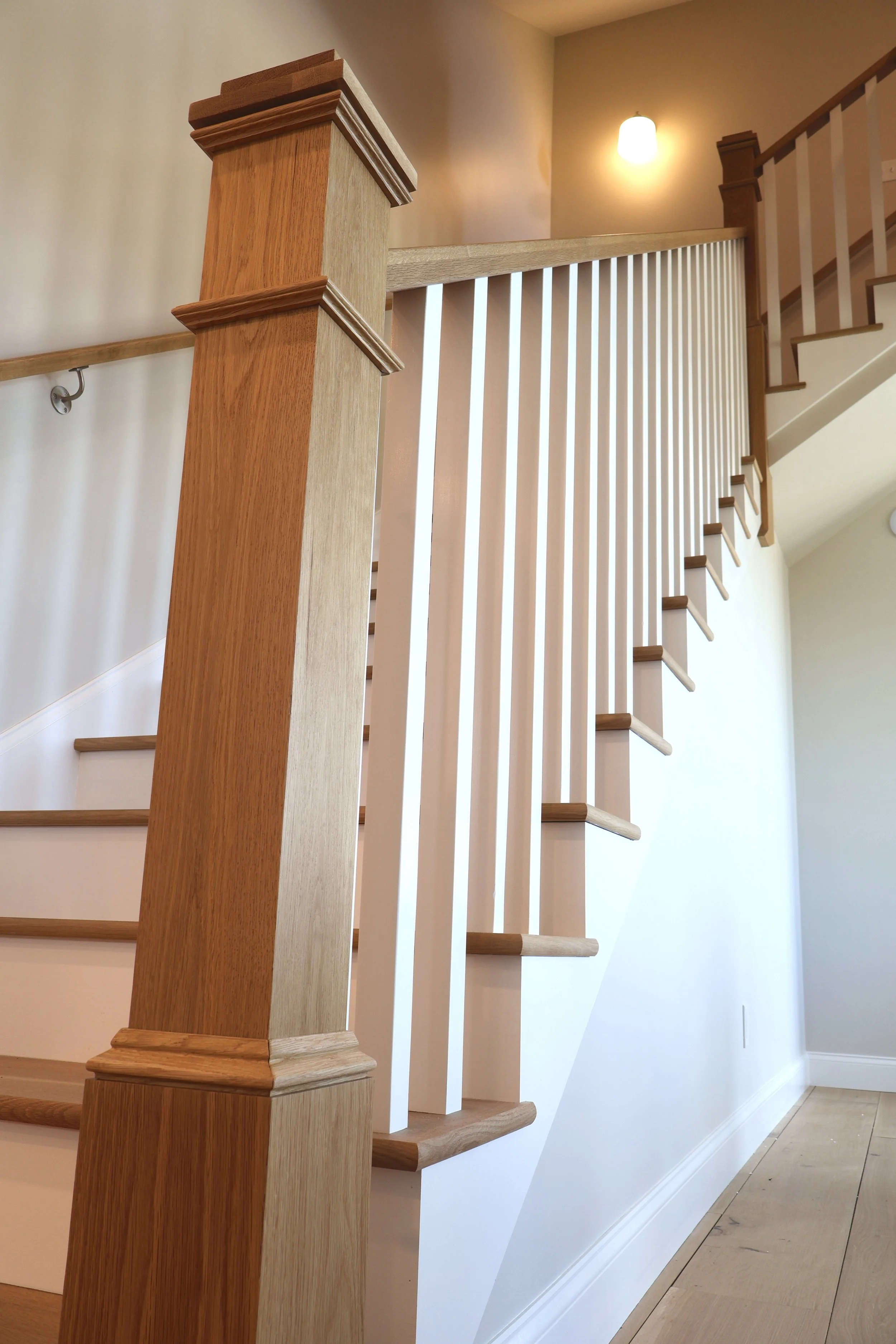 Wooden staircase with white balusters, a wooden handrail, and a wooden newel post at the bottom of the stairs. A wall-mounted light fixture illuminates the upper section of the staircase, with beige walls and a wooden floor at the base.