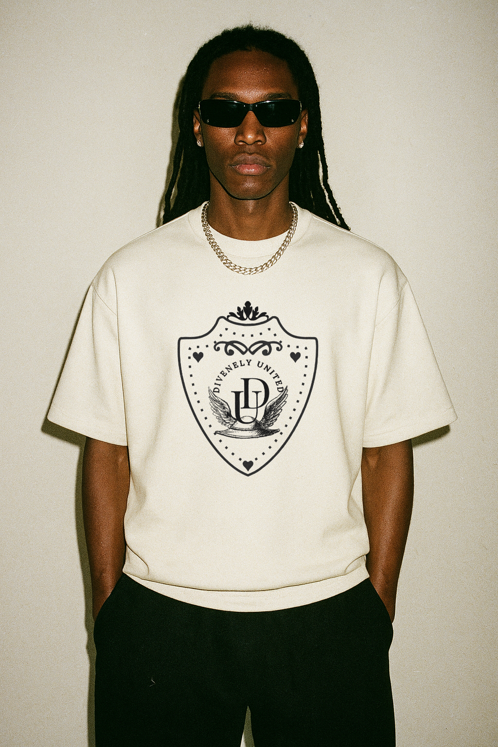 Male model wearing sunglasses, a white DIVENELY UNITED crest T-shirt and black trousers against a light studio background.