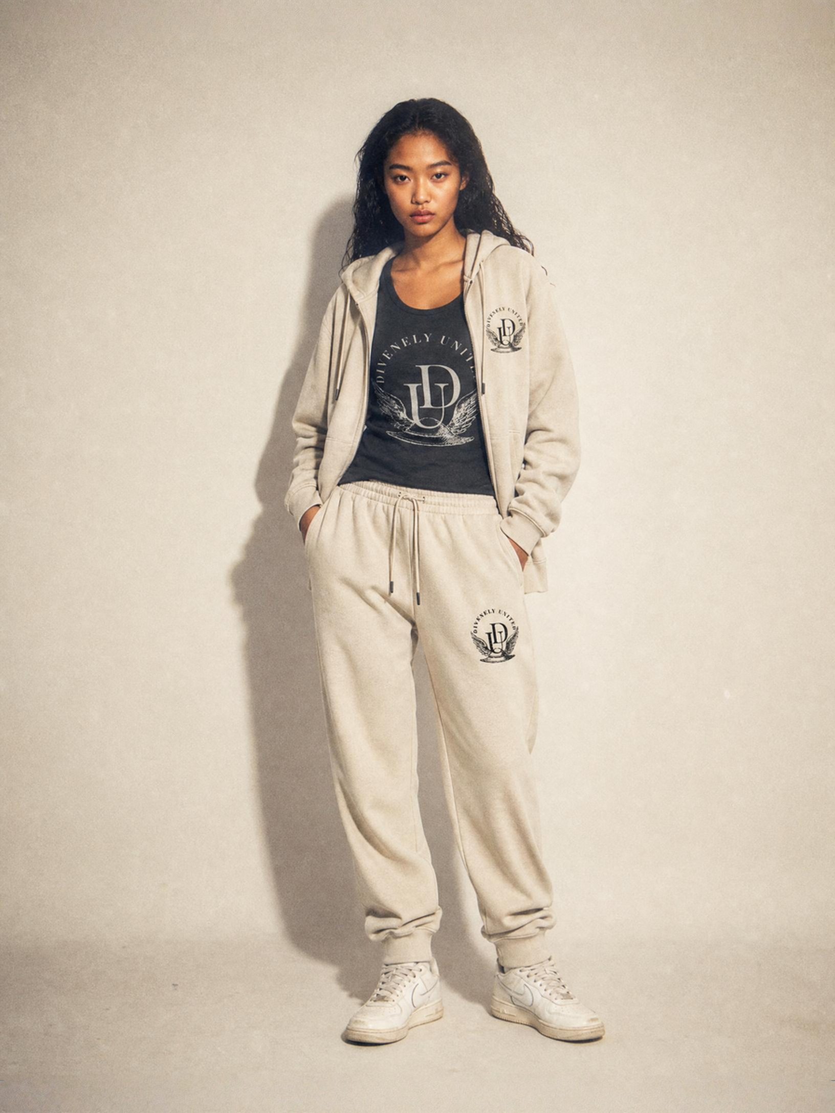 Woman in beige hoodie, matching sweatpants, and white sneakers standing against a plain light-colored wall.
