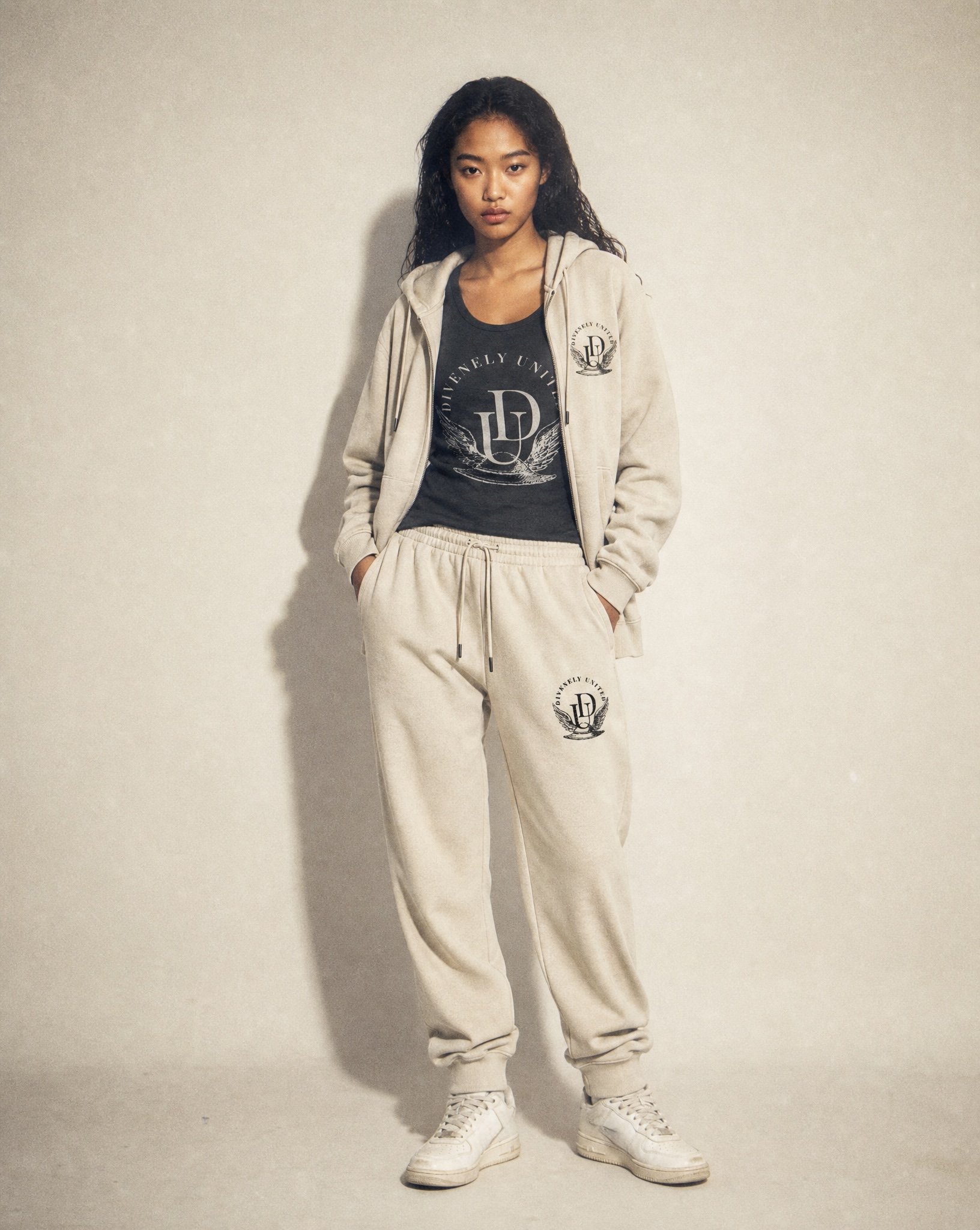 A young woman with dark curly hair wearing a beige hooded sweatshirt with a logo, beige sweatpants with the same logo, white sneakers, and a black t-shirt underneath, standing against a plain off-white background.