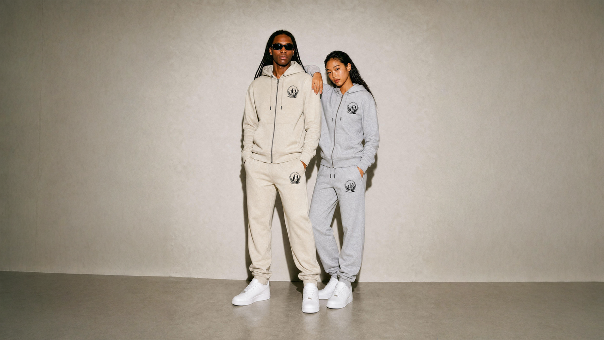 Two models, a male and a female, are standing against a plain beige wall. Both are wearing matching hooded sweatshirts and sweatpants with a logo on the chest and thigh, along with white sneakers. The male model is wearing black sunglasses and has his arm around the female model's shoulder.