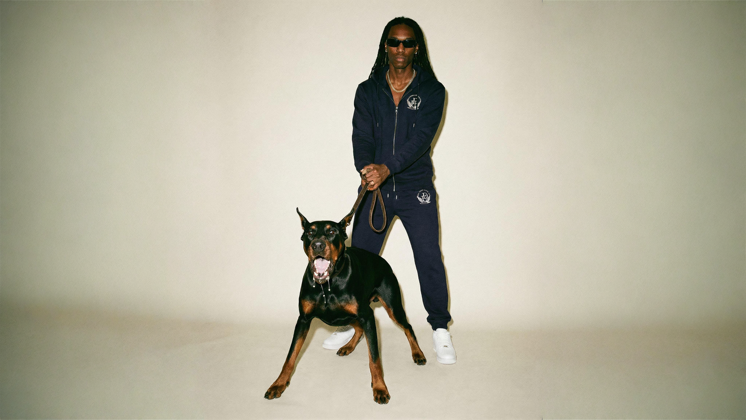 A man wearing a navy blue Divenely United  adjusting his posture and looking down into camera, holding an aggressive drooling leashed doberman barking into camera
