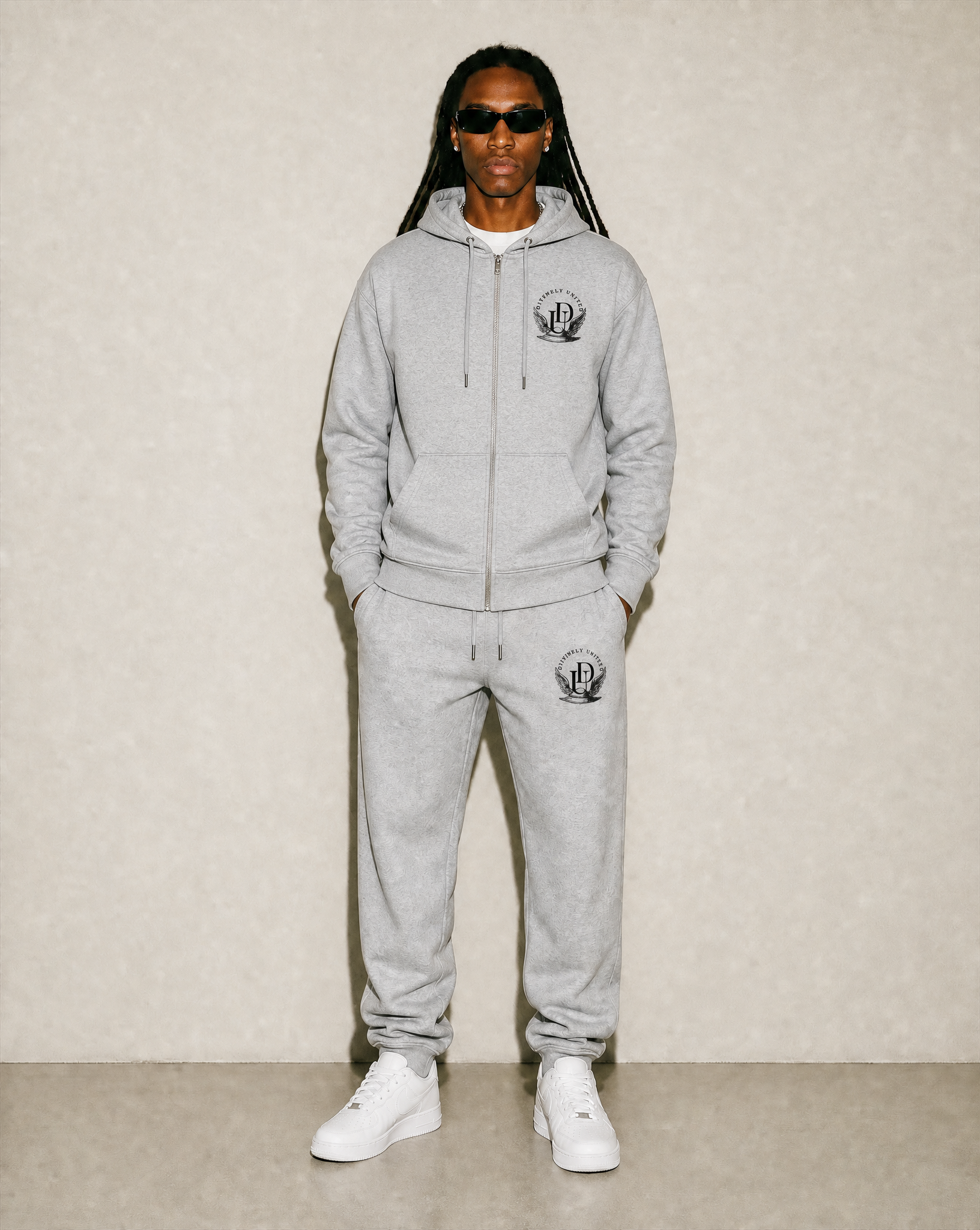 Person standing against a beige wall, wearing a gray hoodie with a logo, matching sweatpants, white sneakers, dark sunglasses, and sporting long dreadlocks.