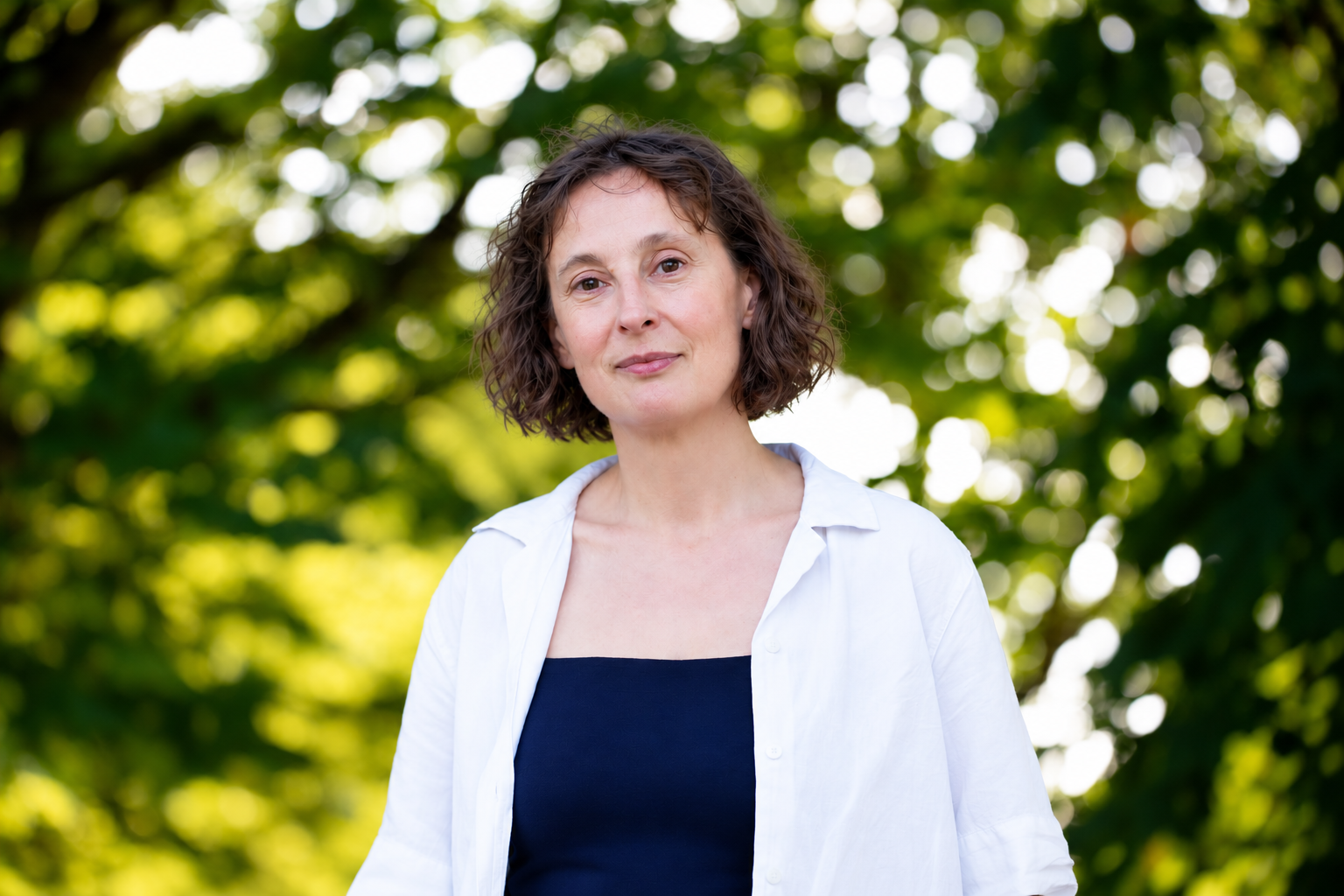 A woman with short curly brown hair standing outdoors in front of green leafy trees, wearing a white shirt over a black top, with a calm expression.