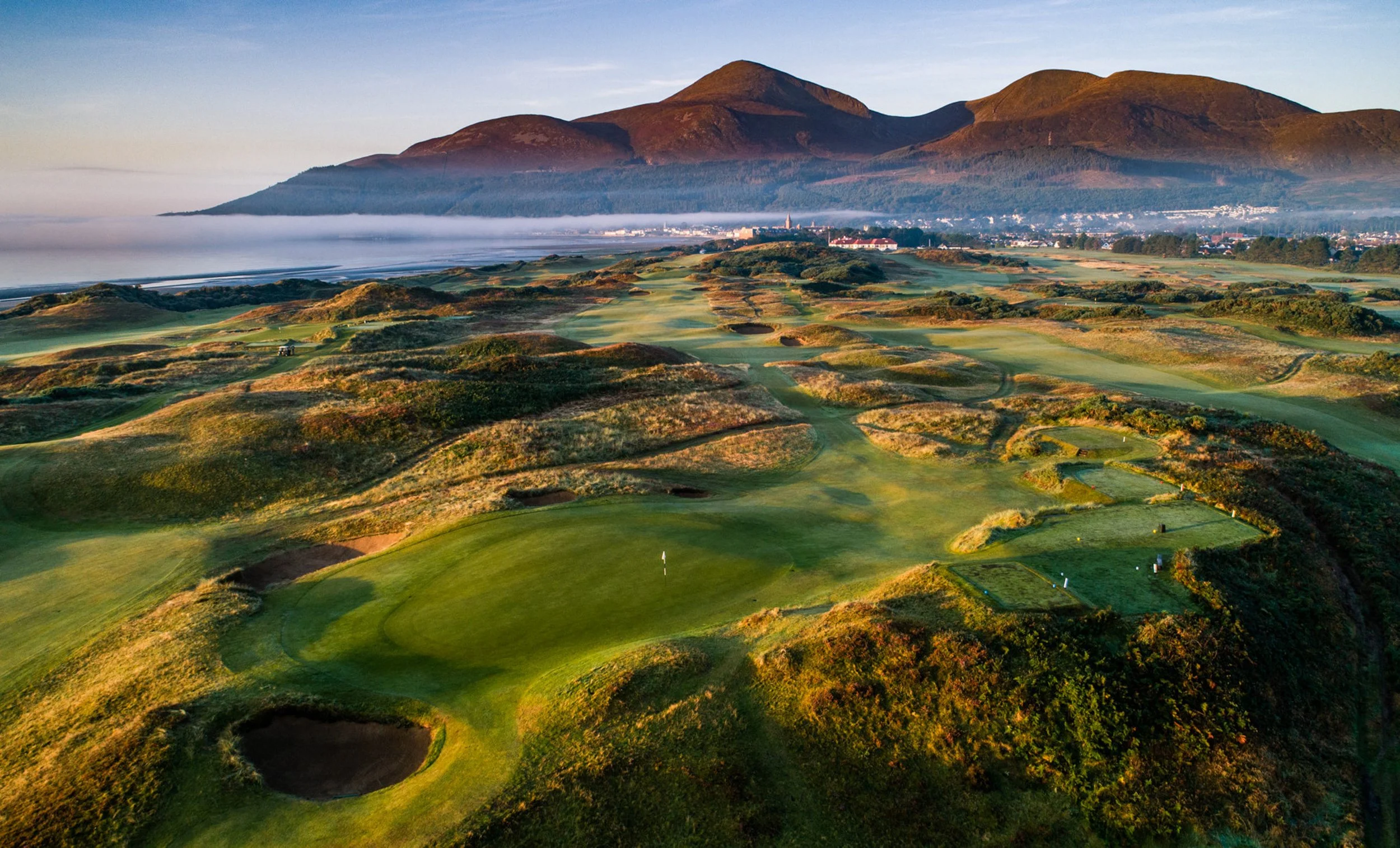 Aerial view of a golf course with green fairways and sand bunkers, surrounded by hills and mountains in the background, during early morning or late afternoon.