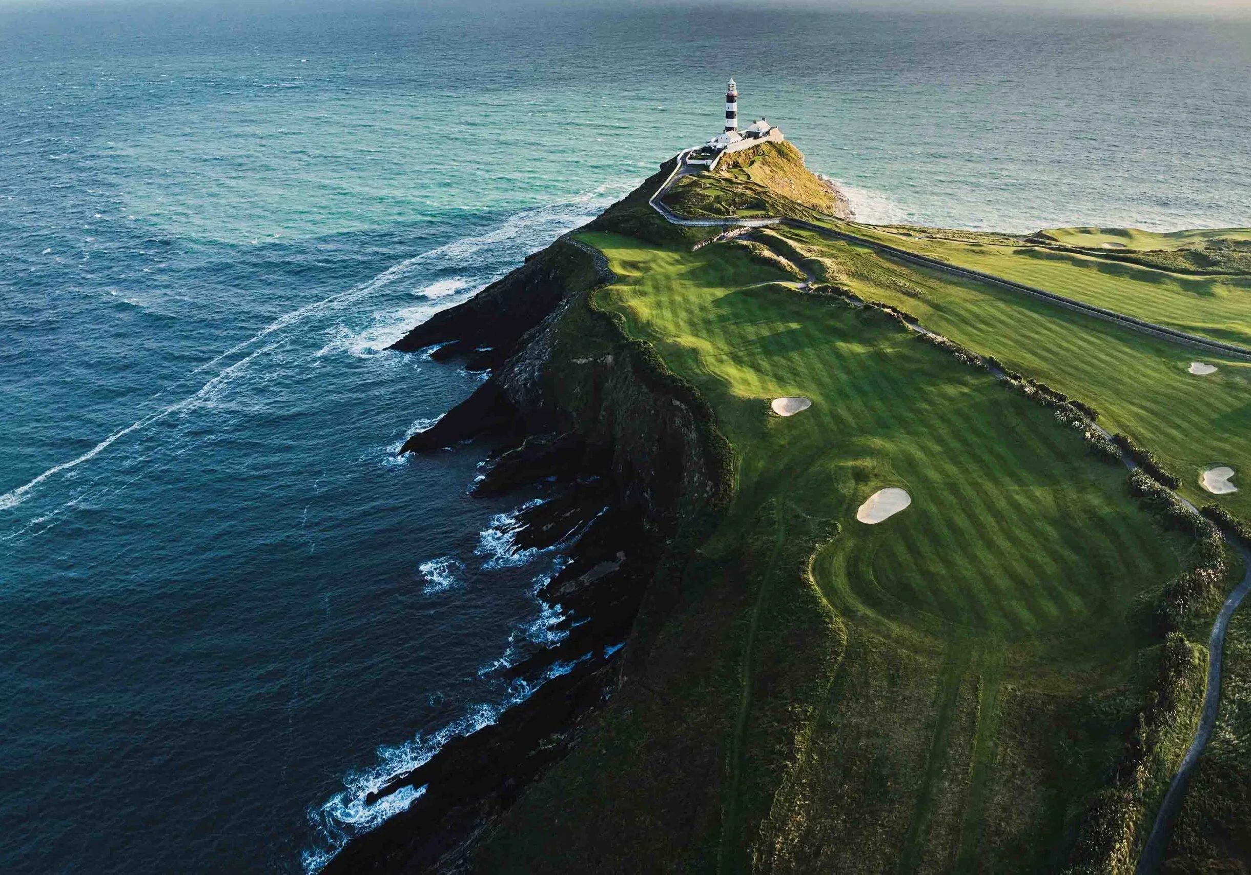 A scenic aerial view of a coastline featuring a lighthouse on a hill, with green golf course and winding roads along the edge of the cliff, overlooking the ocean.