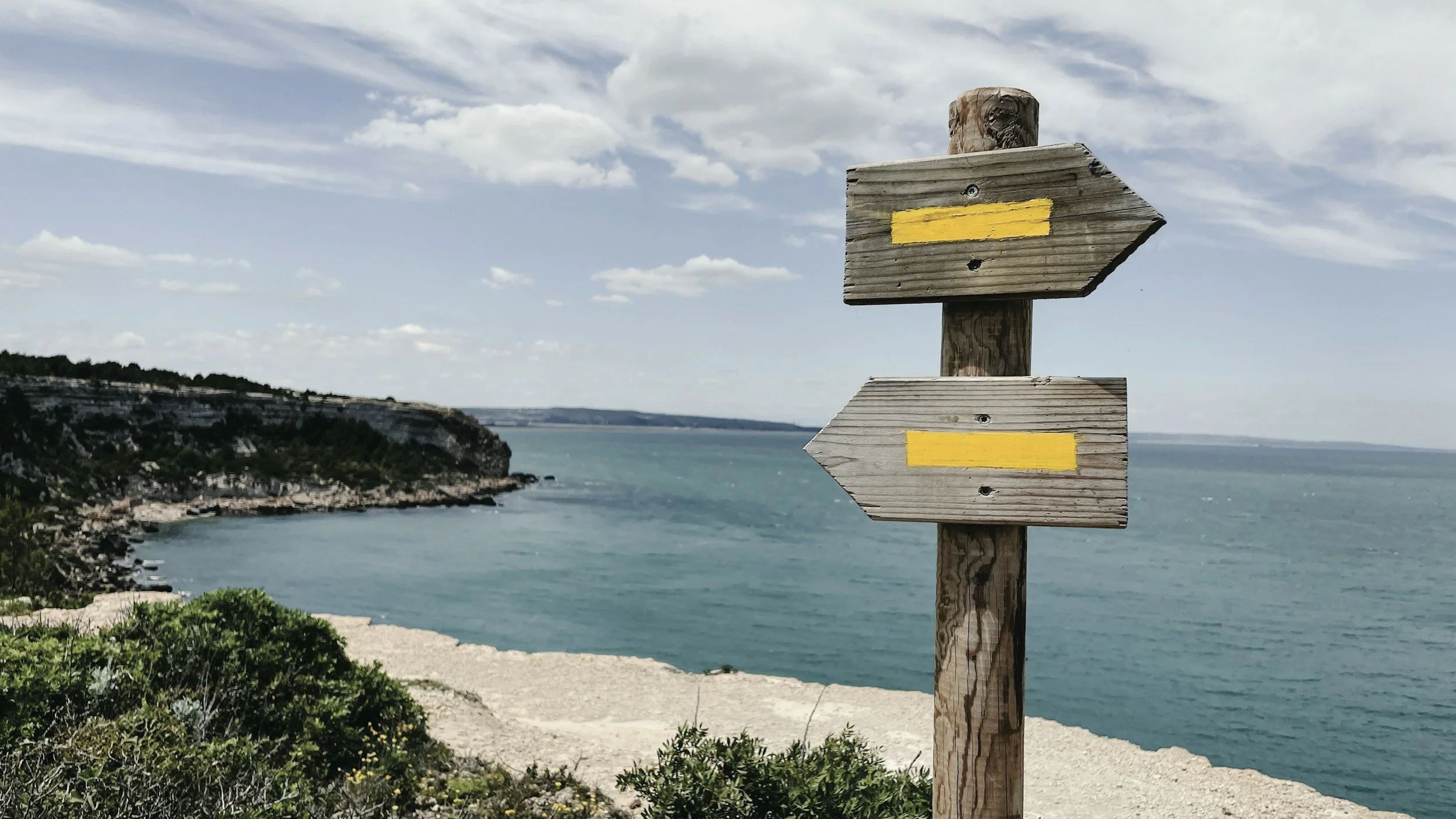 A wooden signpost with yellow markings, situated on a coastal landscape with cliffs, water, and cloudy sky in the background.