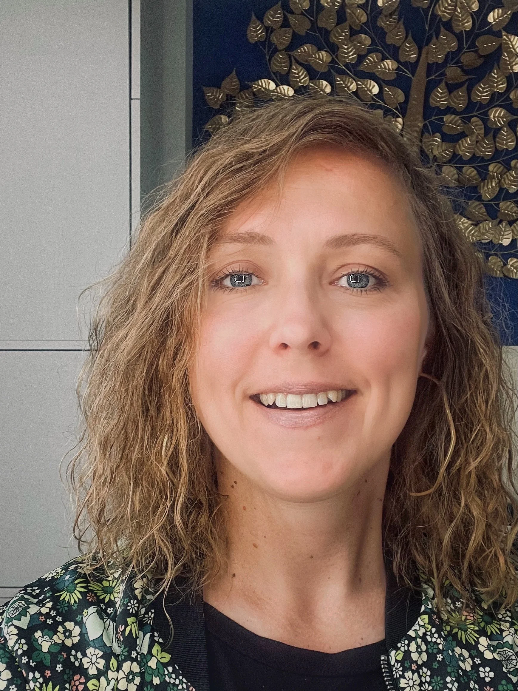 Close-up photo of a smiling woman with curly hair, tan skin, and blue eyes, wearing a floral patterned jacket over a black top, standing in front of a decorative wall with a blue background and gold leaf design.