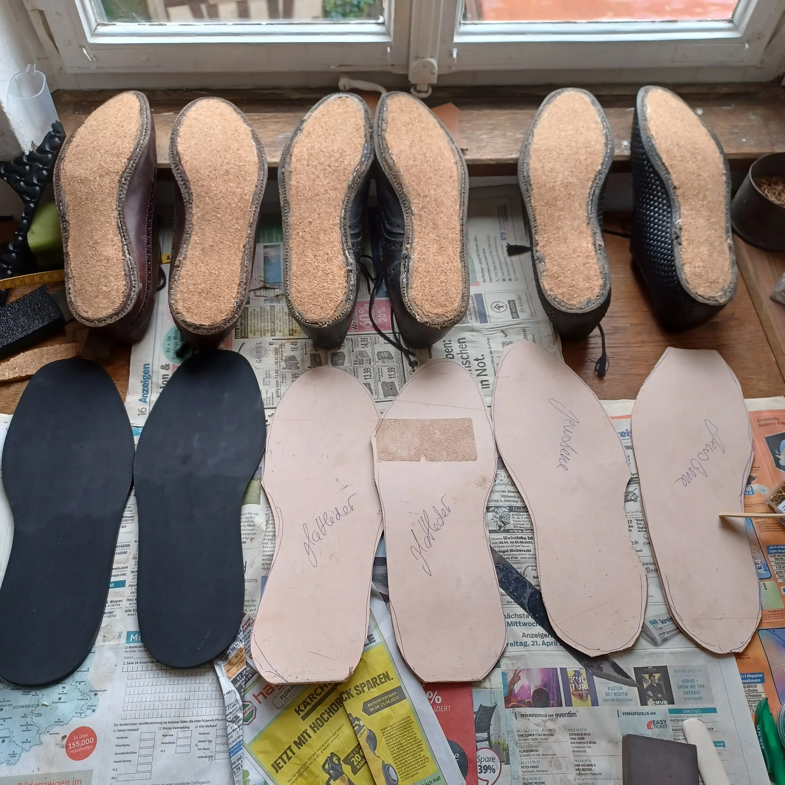 Sohlen von Schuhen, die auf Zeitungspapier vor einem Fenster liegen, einige sind beschriftet und im Verkaufsbereich.