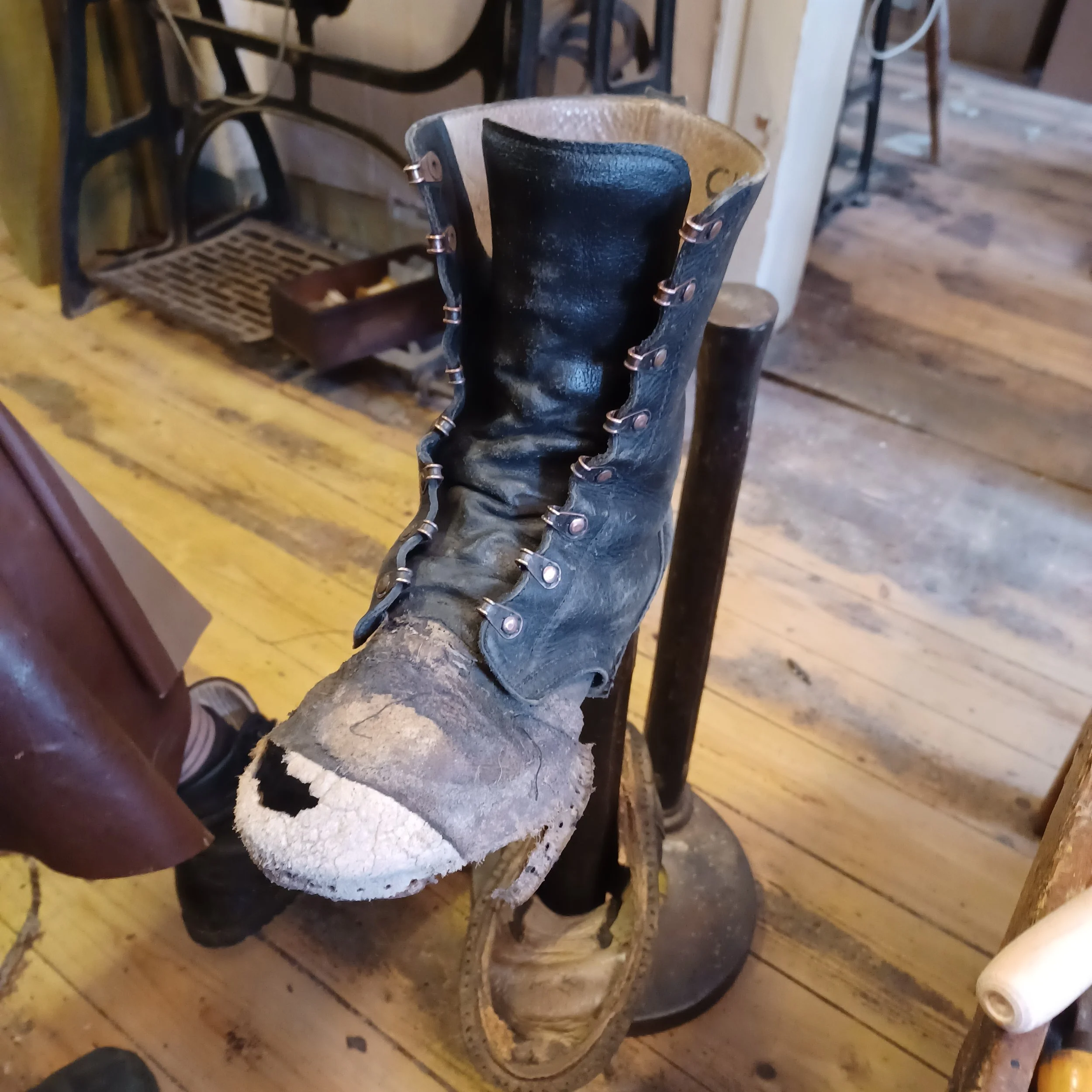 Ein alter, zerbrochener Stiefel mit herausgerissenem Absatz steht auf einem Holztisch. Im Hintergrund sind Teile von Möbeln und eine Holzwand sichtbar.
