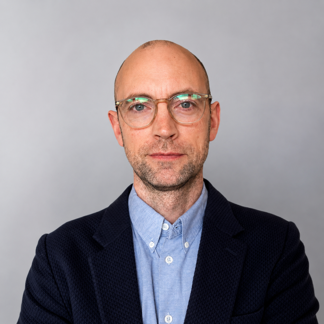 A portrait John Barber, North London Psychotherapist, wearing a dark blazer and a light blue button-up shirt, against a plain gray background.