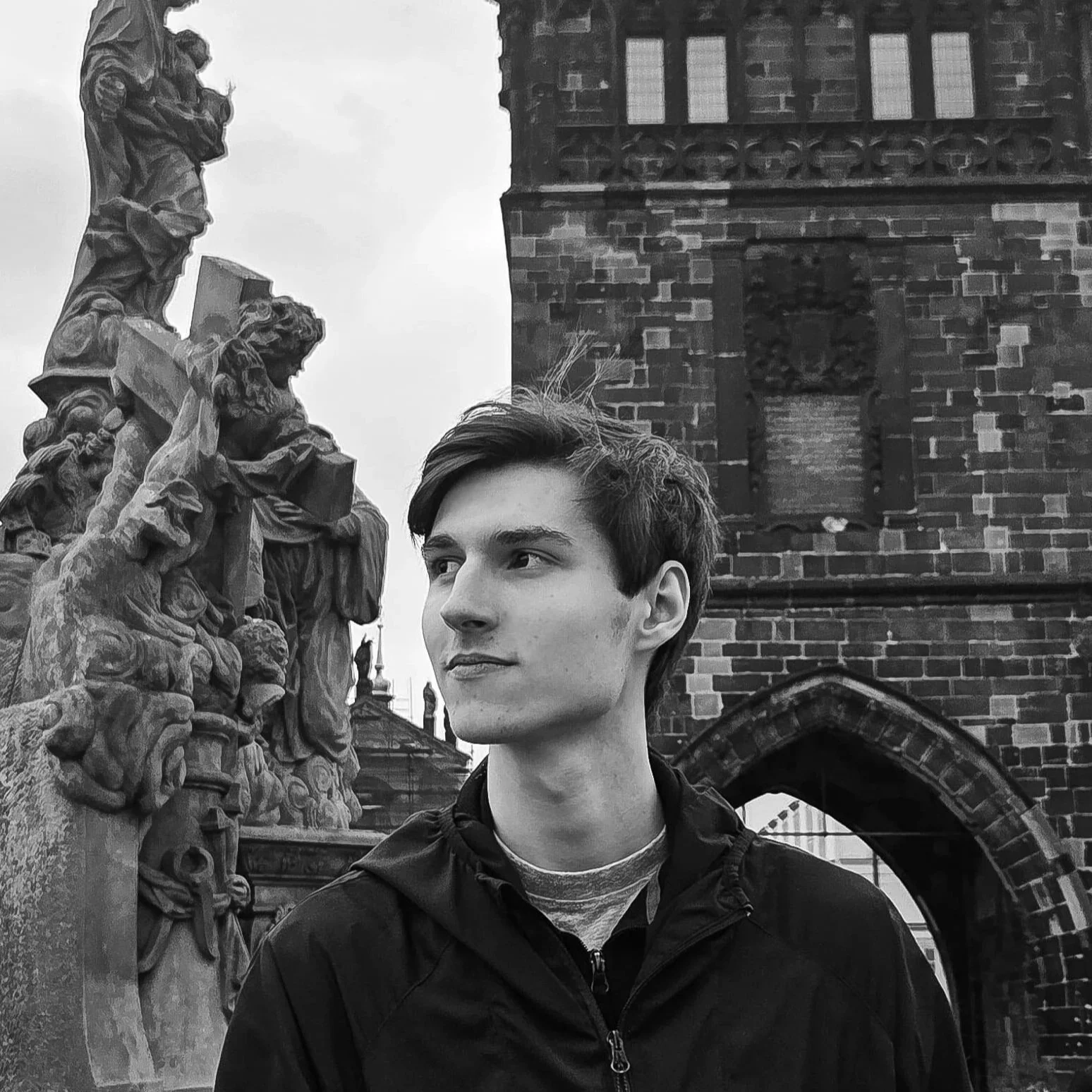 A black-and-white photo of a young man with light skin and short brown hair, standing outdoors near a stone sculpture and a brick building with arched window. The young man is wearing a dark jacket and looking slightly to the right.