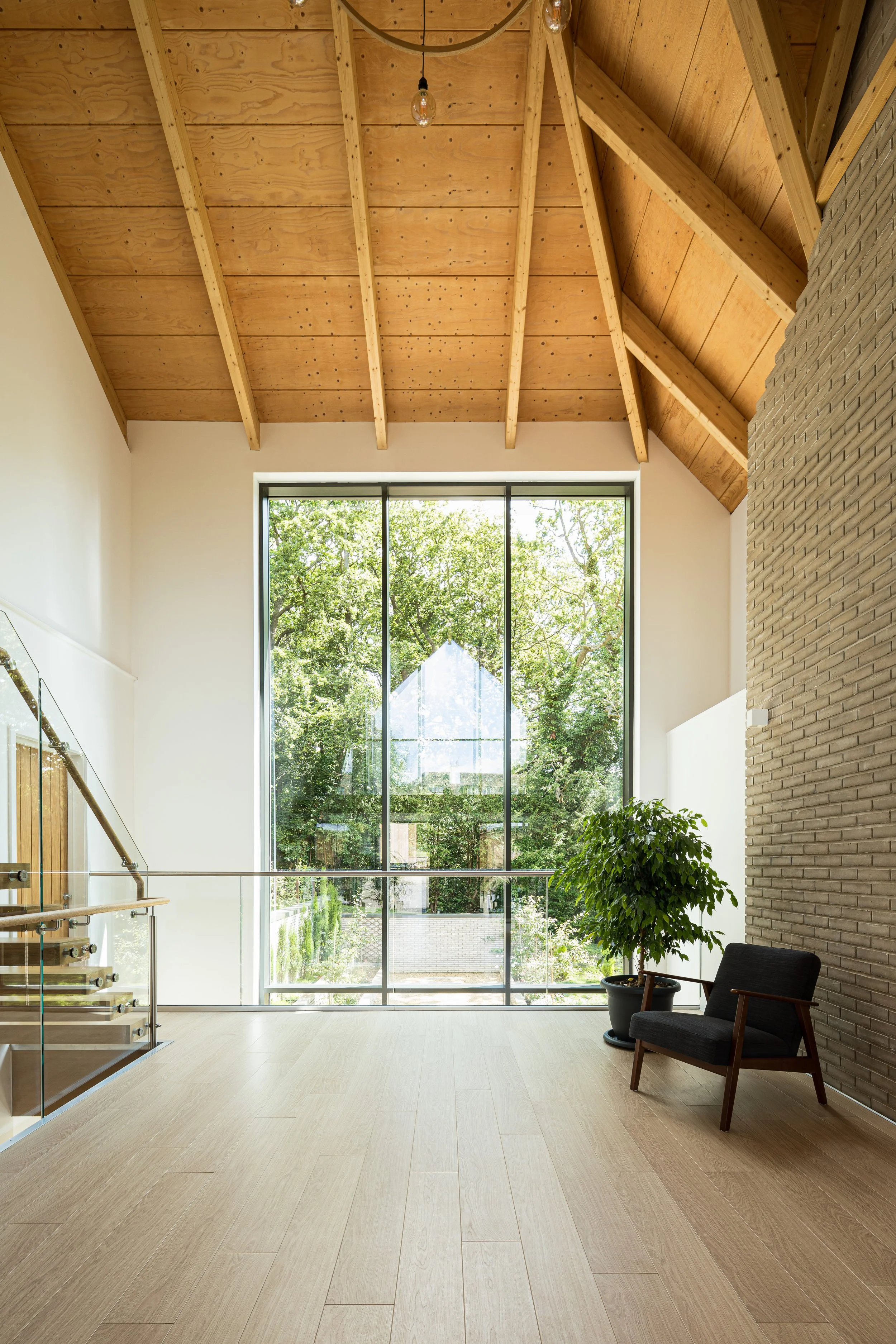 Levitate Architects' Highoaks: An upper-level room with an armchair positioned in front of a striking pointed gabled window that frames a lush green forest view.