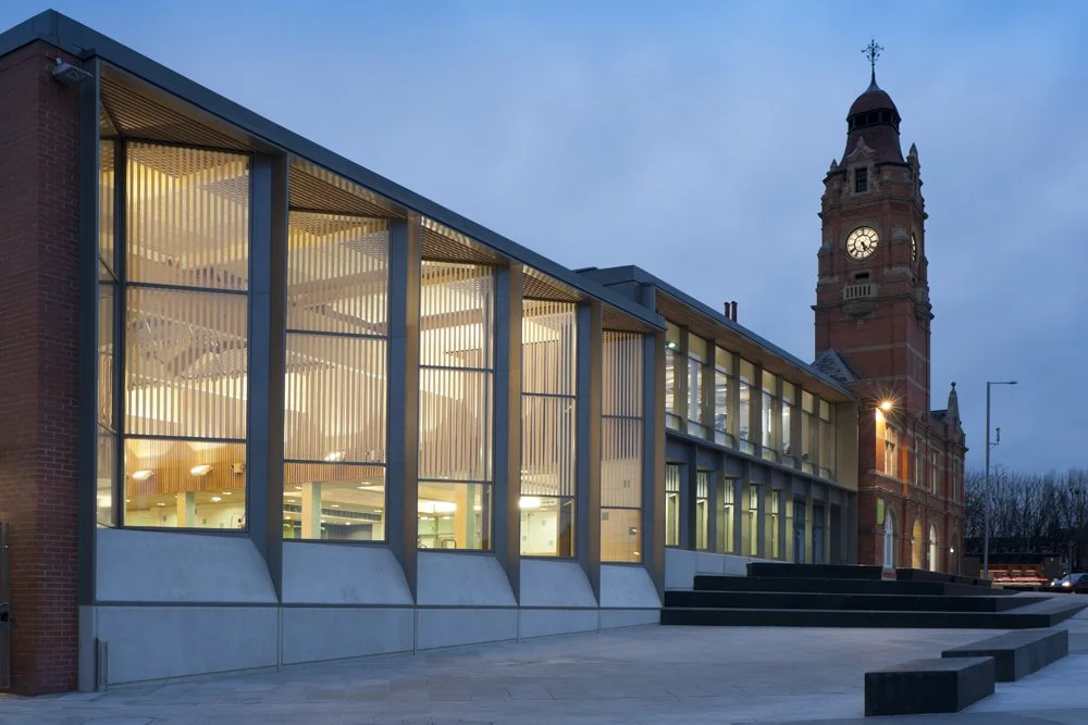 Levitate Architects' Victoria Leisure Centre: The modern glass-fronted exterior illuminated at dusk, showcasing the indoor pool area next to the historic brick clock tower.