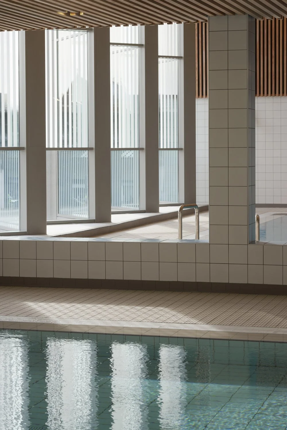 Levitate Architects' Victoria Leisure Centre: A close-up architectural detail of the clean white poolside tiling, reflection in the water, and vertical timber slat walls near the windows.